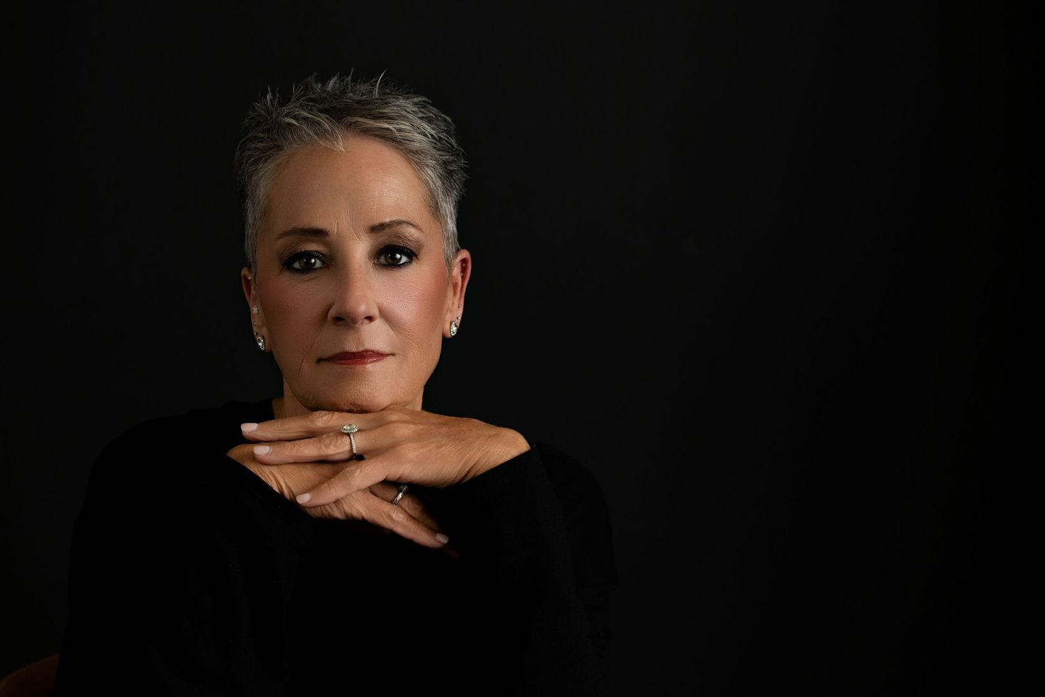 A headshot  by Melissa Nansen Photography short gray hair in a black outfit, resting chin on clasped hands against a dark backdrop.