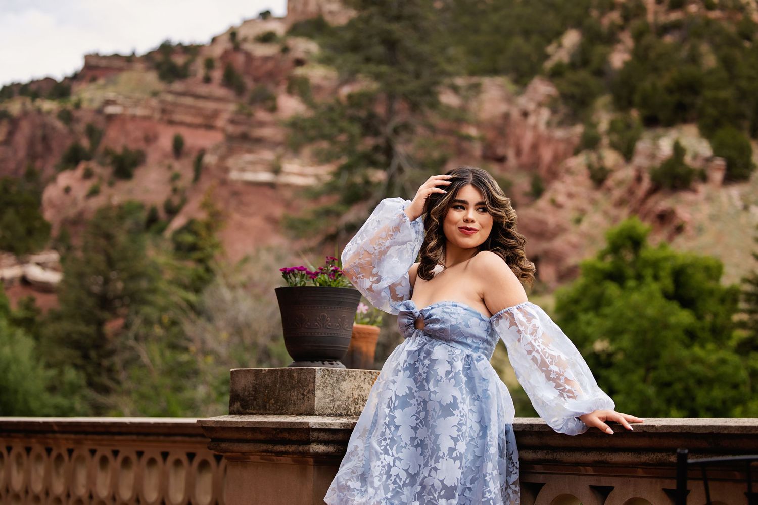 Young woman in blue off-shoulder lace dress posing on stone balcony with red rock mountain backdrop for Littleton Colorado Senior pictures