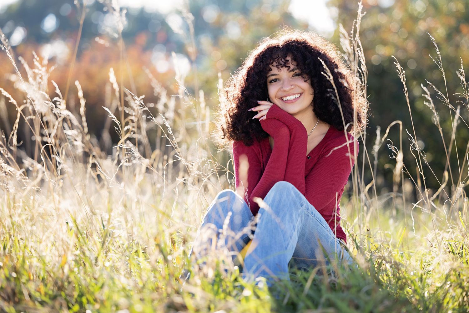 A young woman with curly hair sits smiling in a sunlit field of tall grass, wearing a red top and jeans Aurora Colorado
