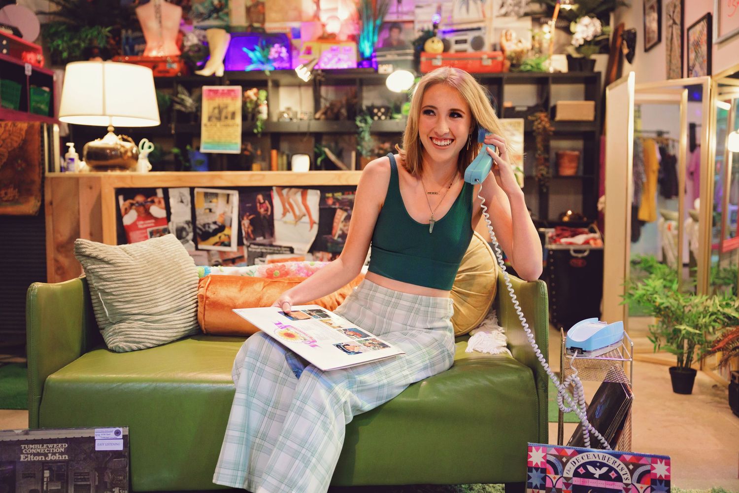 Smiling woman sitting on green sofa talking on retro phone, holding magazine in colorful eclectic shop in Denver thrift shop