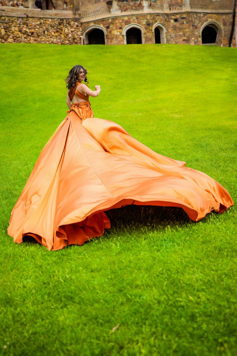 High School girl spinning in voluminous orange ballgown on green grass lawn beside stone castle archways for senior photos in the springs