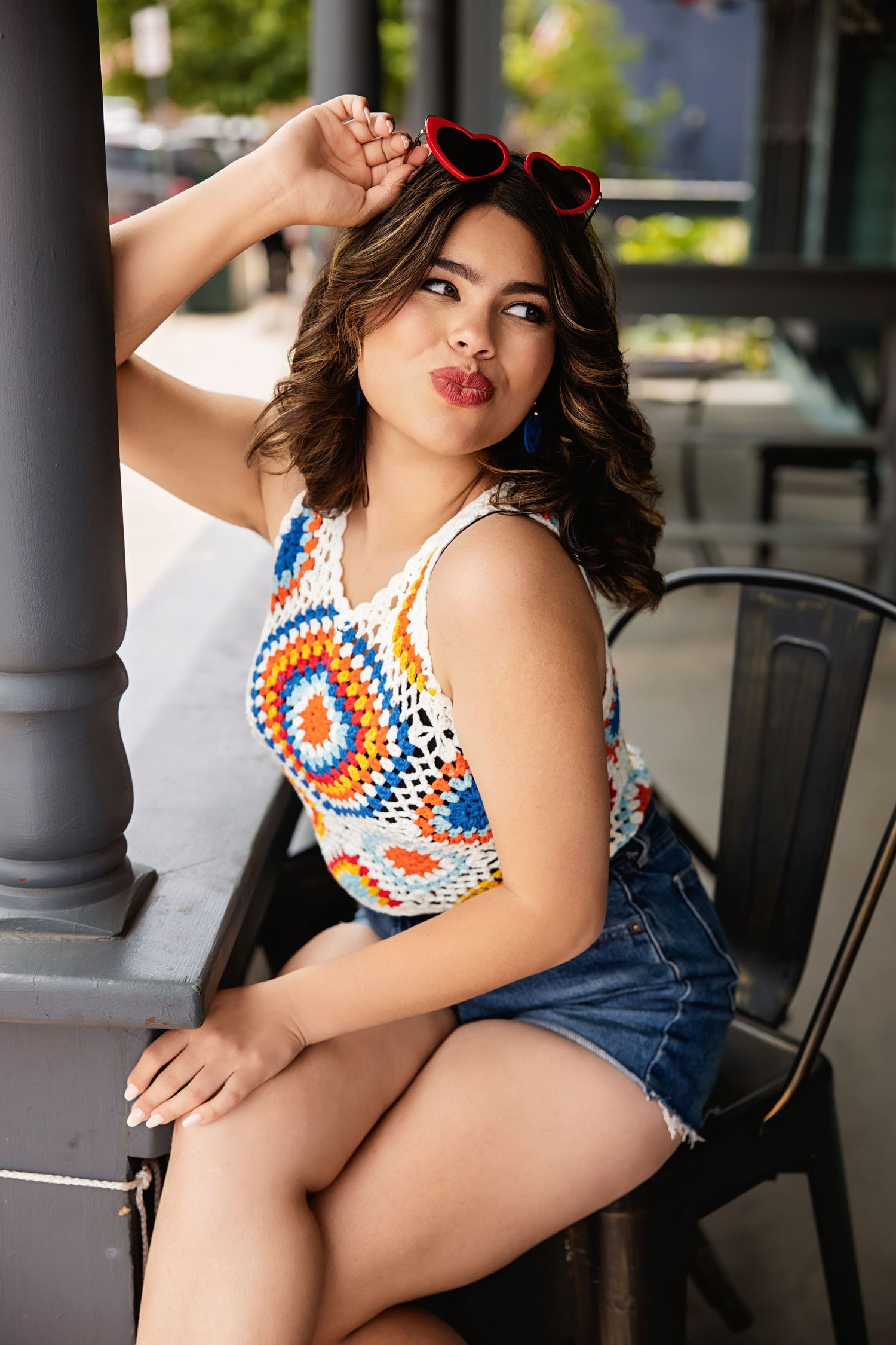 Fashionable woman in multicolor crochet top with red heart sunglasses sitting at outdoor cafe for Parker CO High School Senior pictures