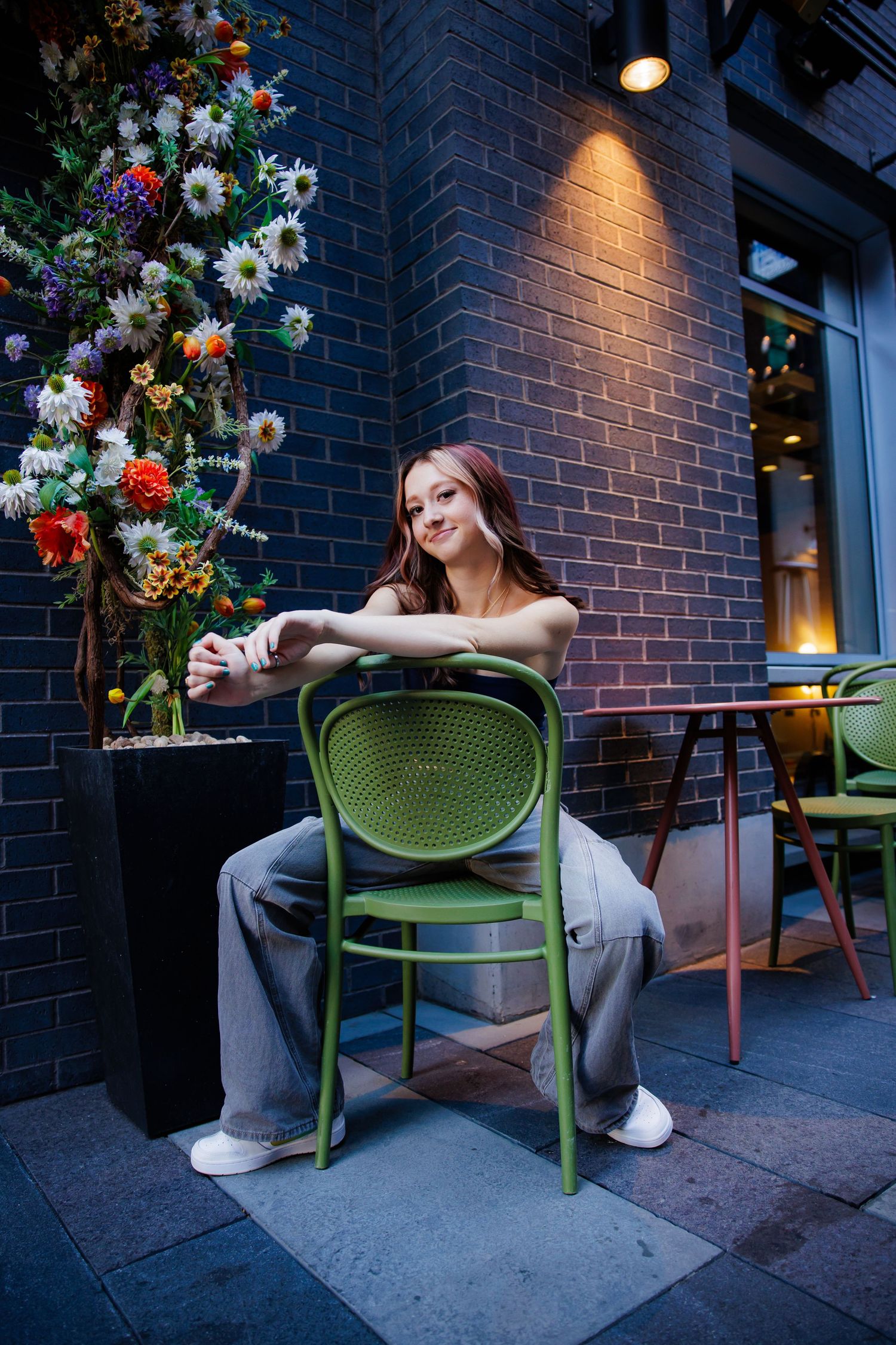 Young woman with pink hair sitting backwards on green chair at outdoor cafe with flowers for Highlands Ranch Colorado Senior pictures