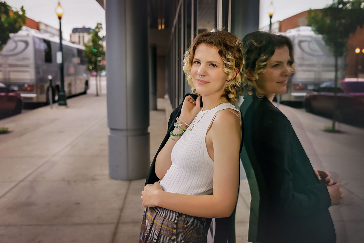 Smiling curly blonde in white tank top leaning against reflective glass pillar on city sidewalk.