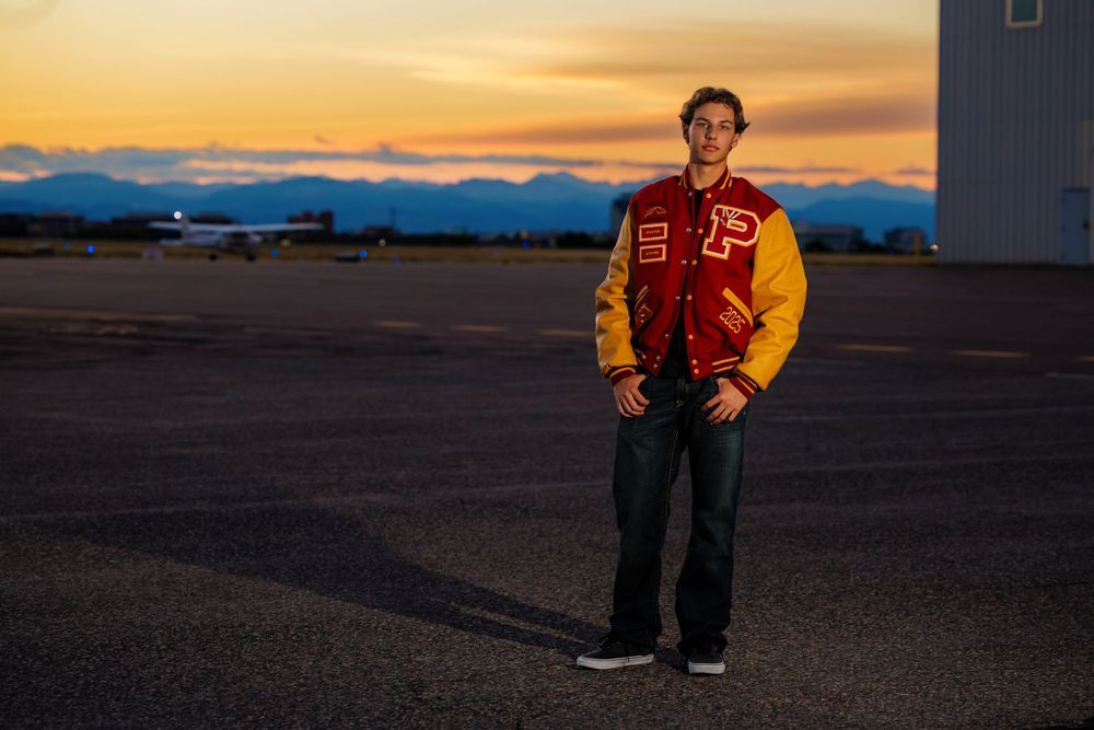Teen in red and gold varsity jacket stands on tarmac during colorful evening sunset. Lonetree Colorado Senior Portrait