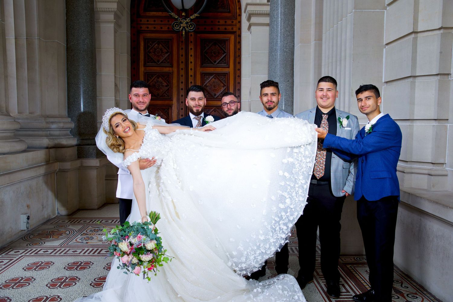 Wedding party lifting bride's gown as groom and groomsmen pose at ornate building entrance.