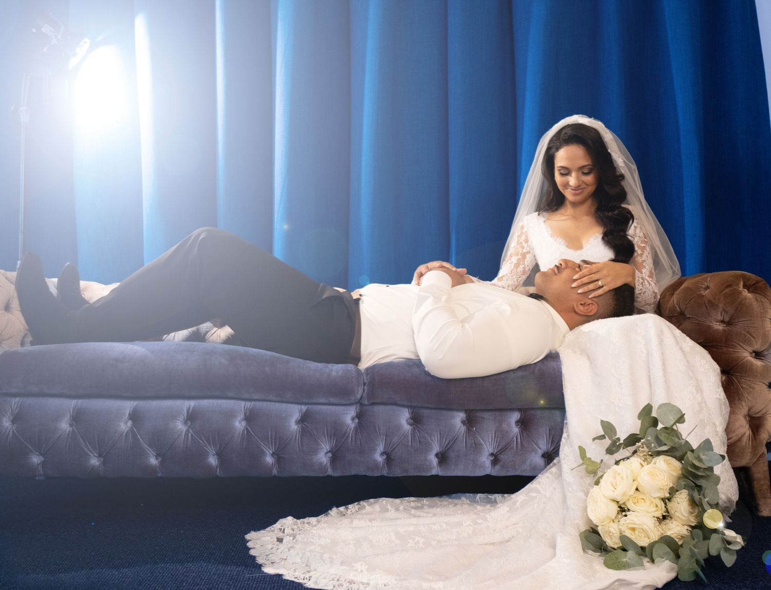 Elegant bride in white gown resting on a velvet sofa with a bouquet of white roses nearby.