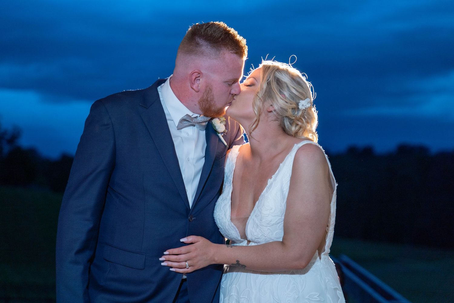 Bride and groom share a tender kiss at dusk under a dramatic blue evening sky outdoors.