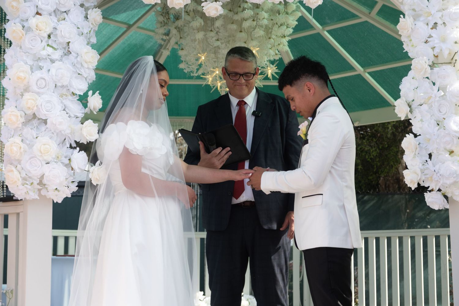 Groom places wedding ring on bride's finger during outdoor ceremony under white floral arch.