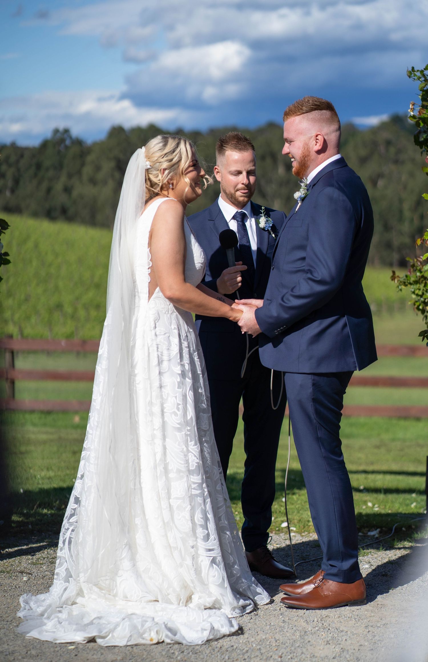 Bride and groom exchange vows holding hands at outdoor vineyard wedding ceremony with officiant.