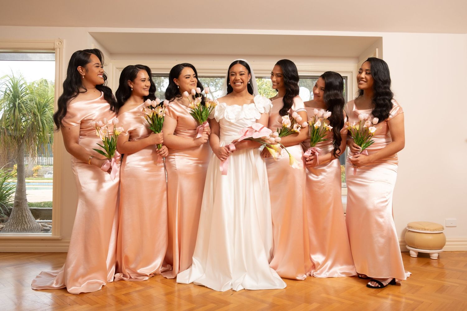 Bride in white dress laughs with five bridesmaids in blush gowns holding tulip bouquets.