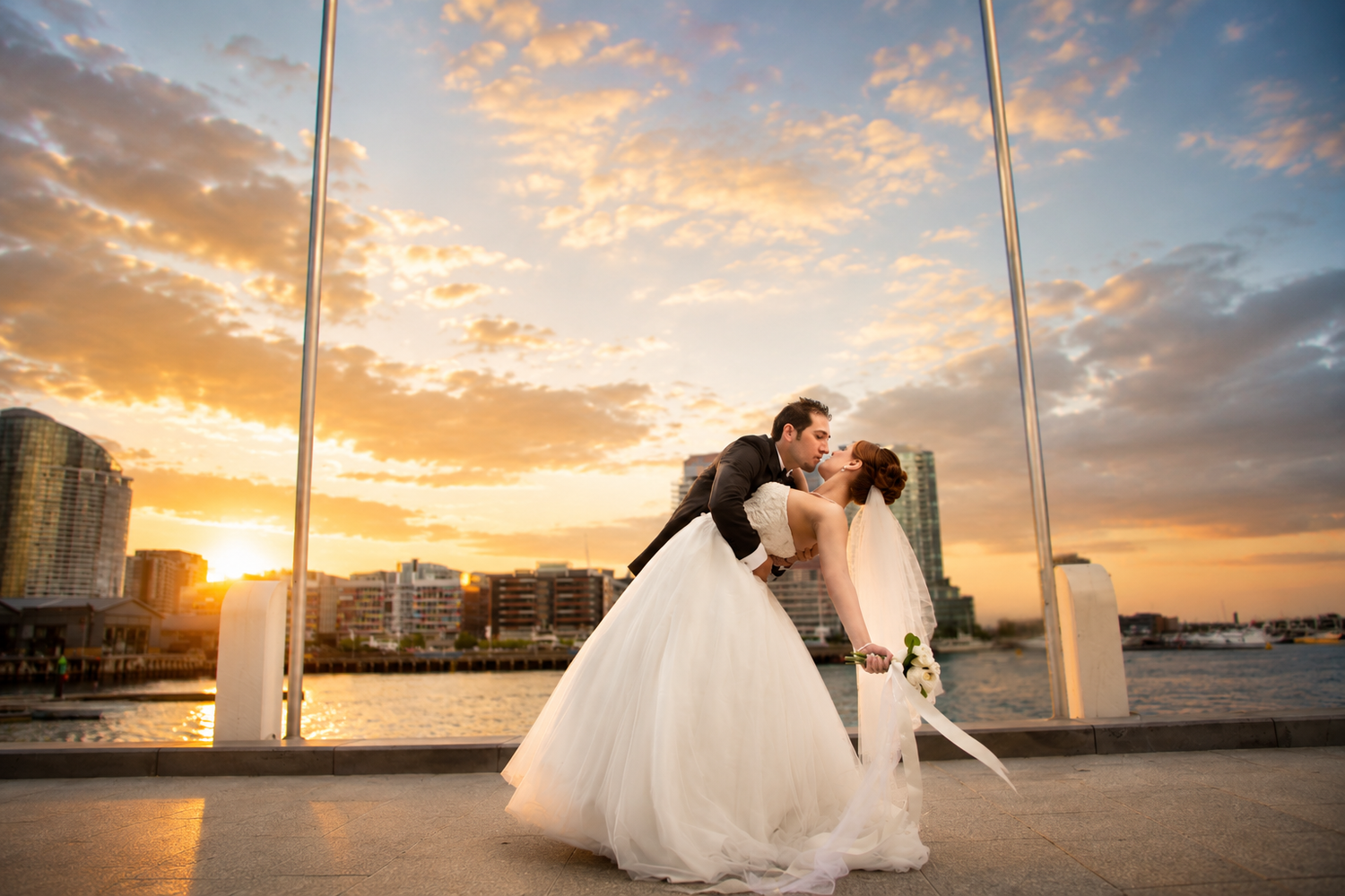 Couple shares a romantic dip kiss at waterfront sunset wedding venue with city skyline backdrop.