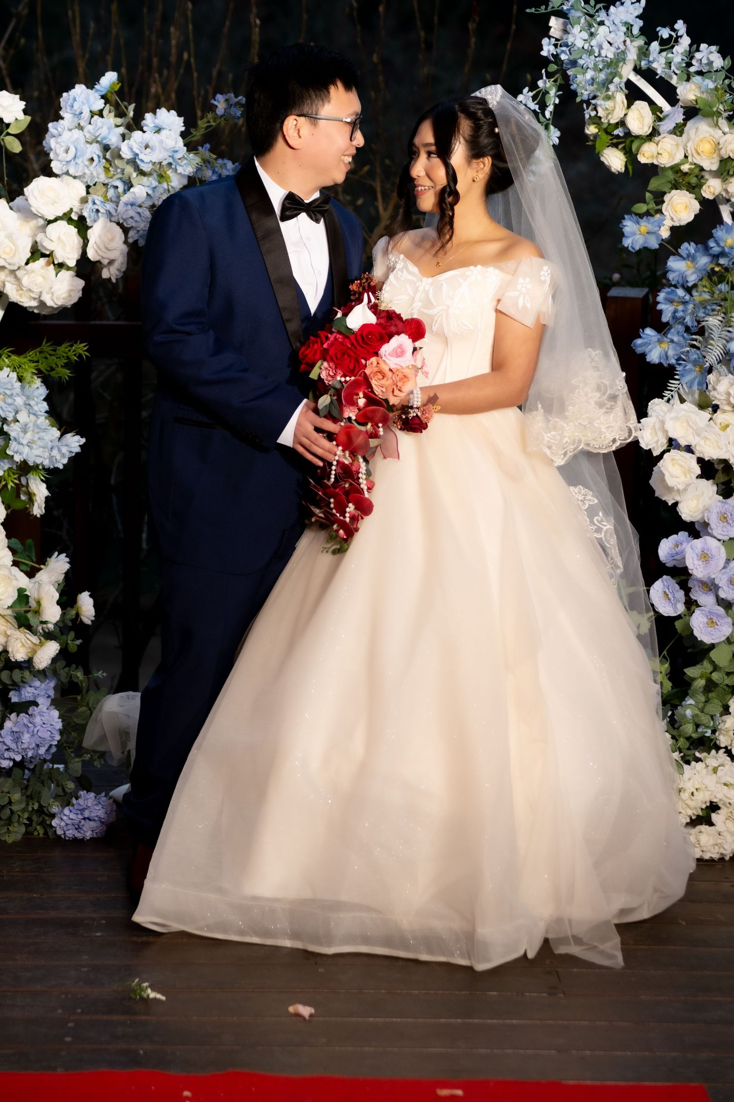 Bride and groom standing together before blue and white floral arch, bride holding red cascading bridal bouquet.