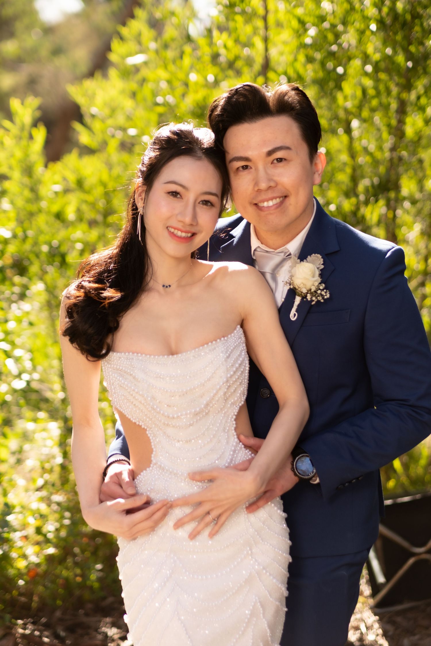 Smiling Asian bride and groom posing together in a lush green garden on their wedding day.
