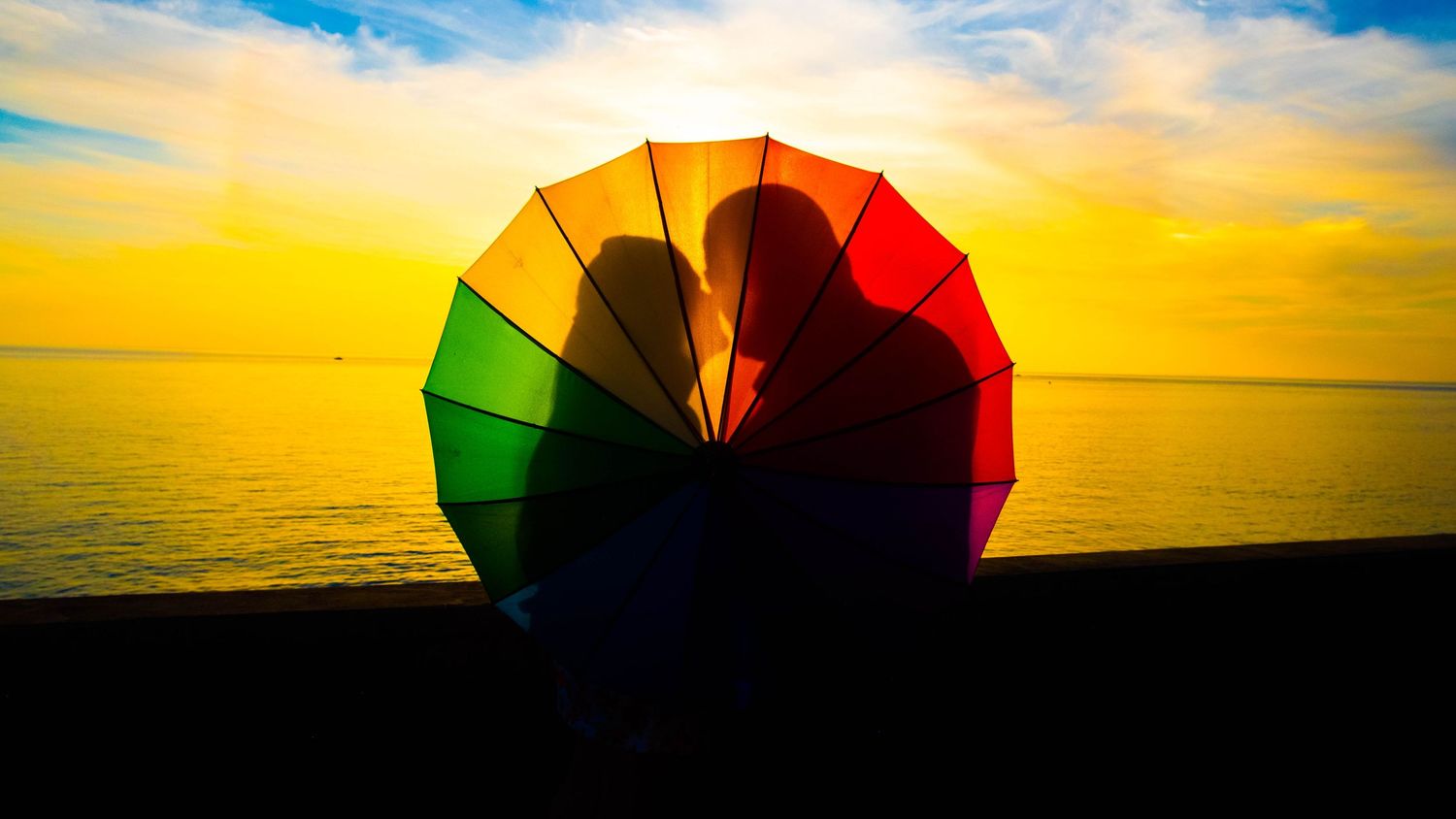 Couple silhouetted behind colorful rainbow umbrella at sunset over ocean horizon.