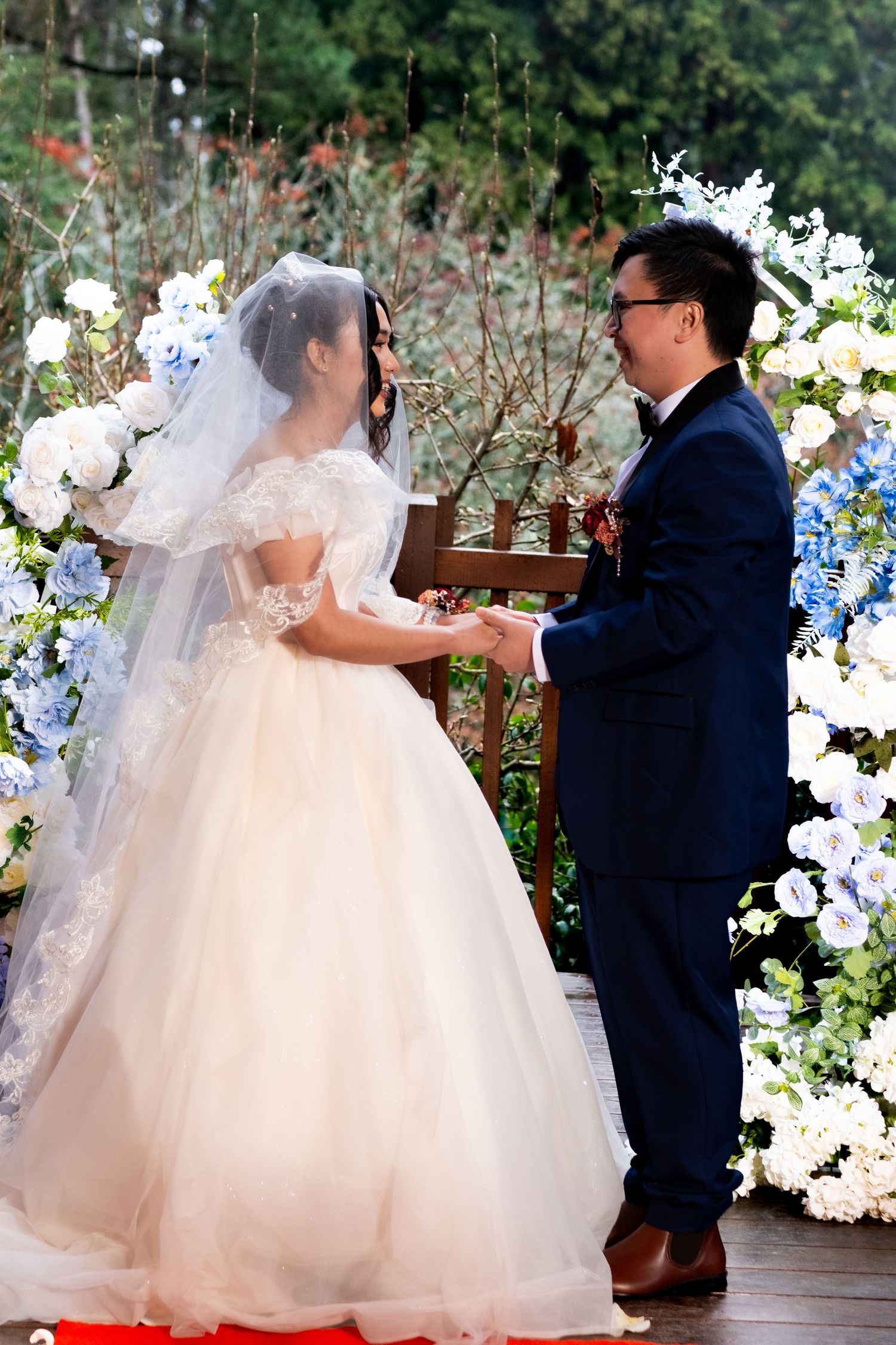 Bride in white ball gown and groom in navy suit holding hands at an outdoor ceremony with blue floral arch.