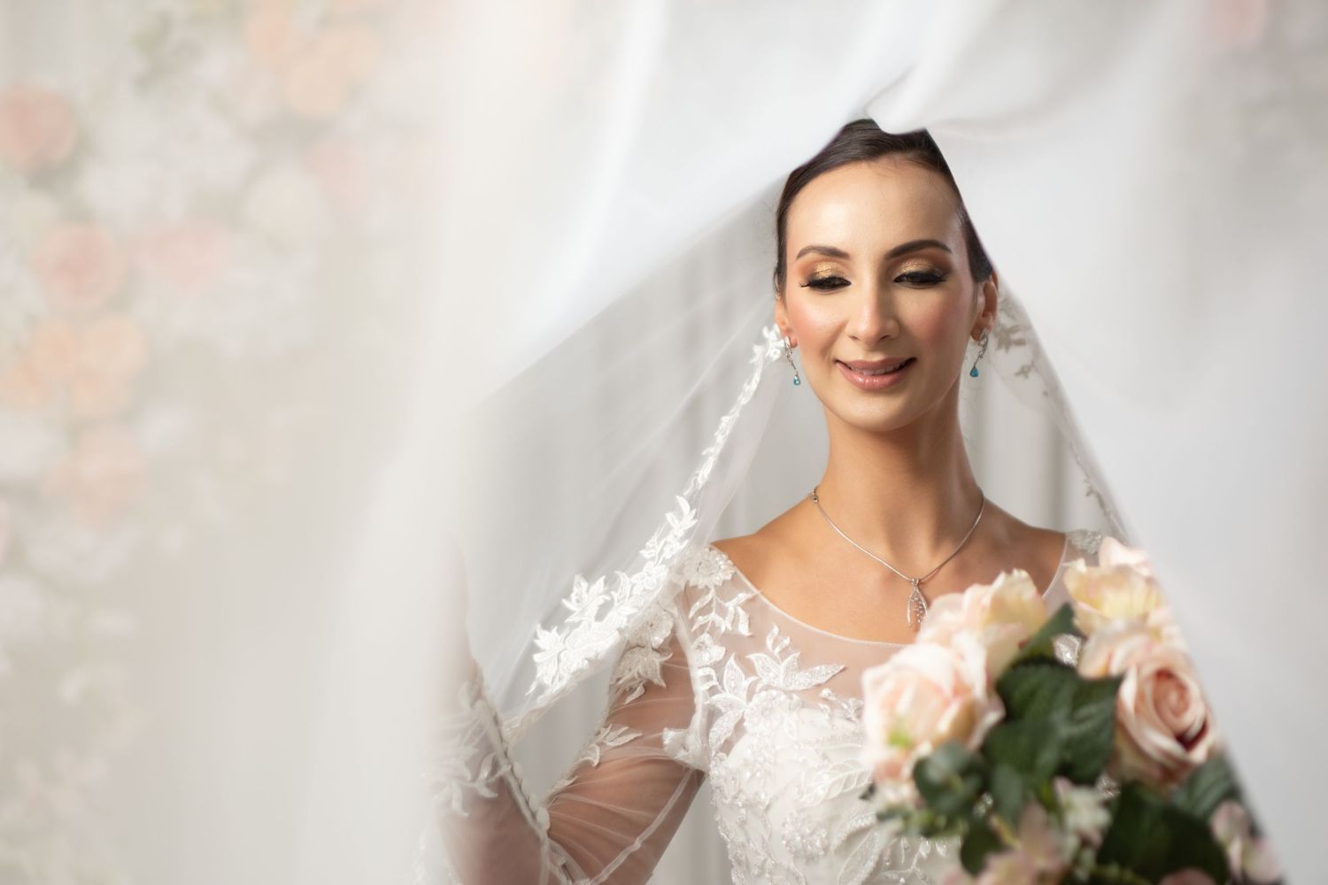 Elegant bride in lace wedding gown holding pink roses bouquet, smiling softly beneath a flowing white veil.