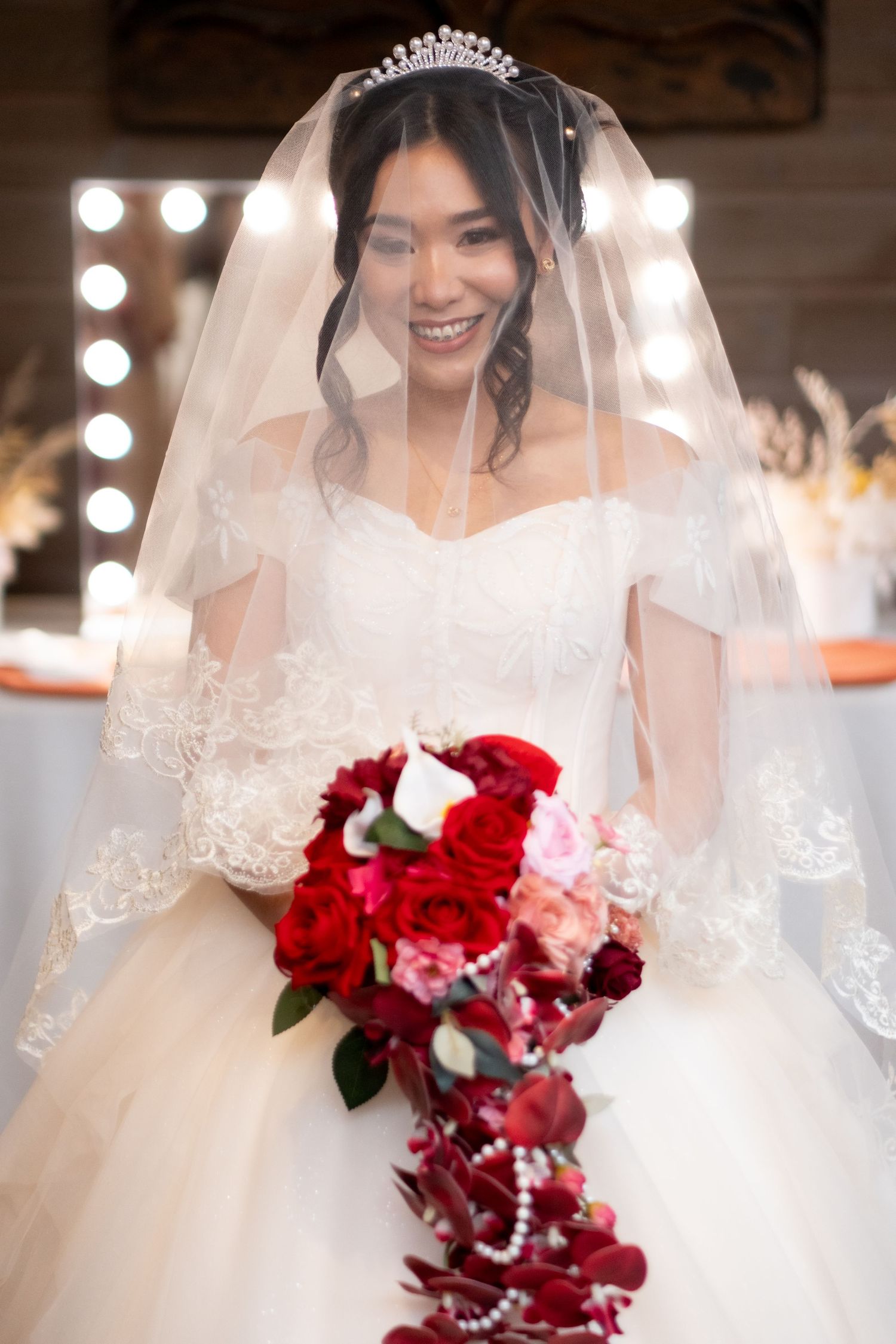 Smiling Asian bride in white lace gown and tiara holds cascading red rose bouquet with veil draped over her face.