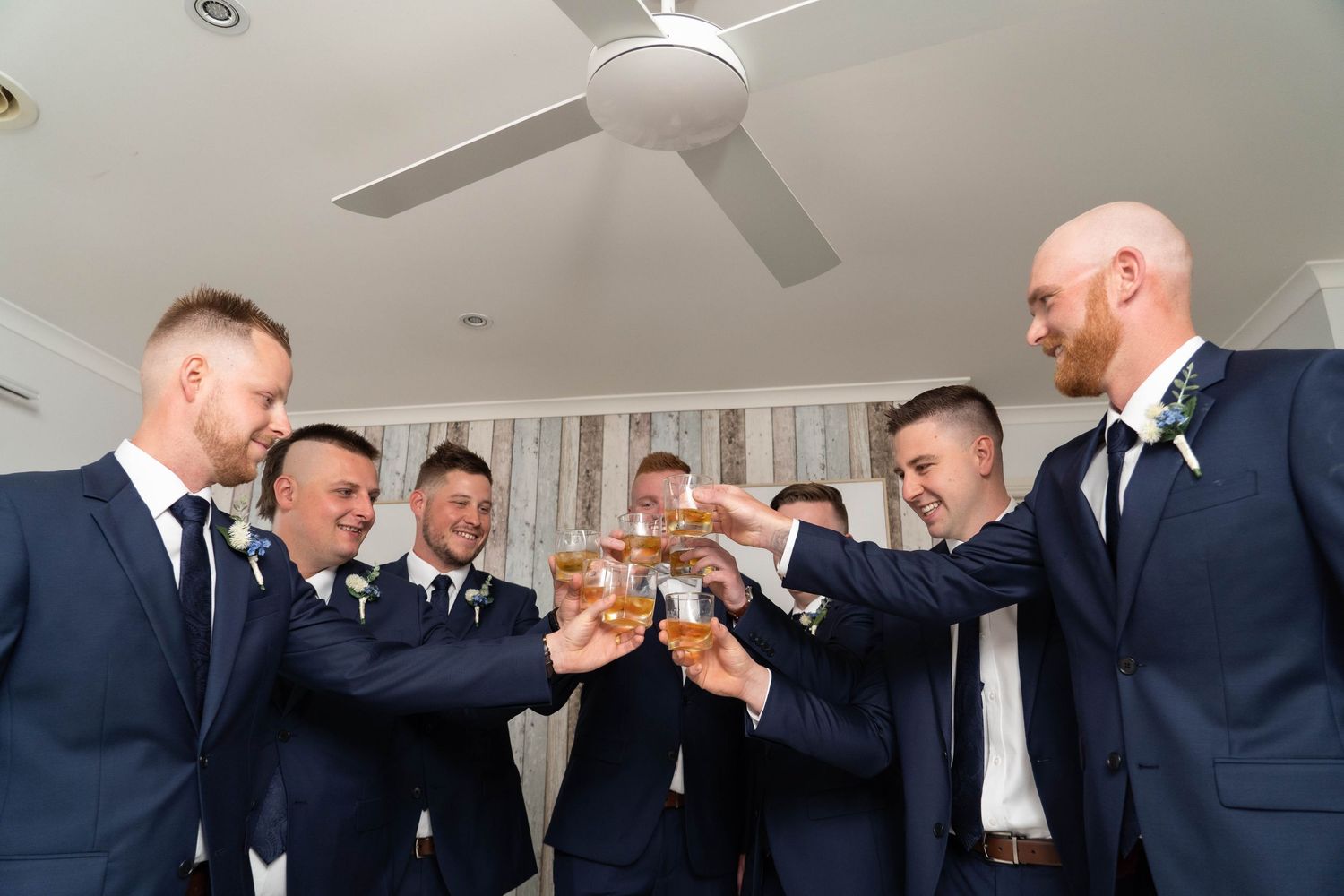 Groom and groomsmen in navy suits toast with drinks in bright room.