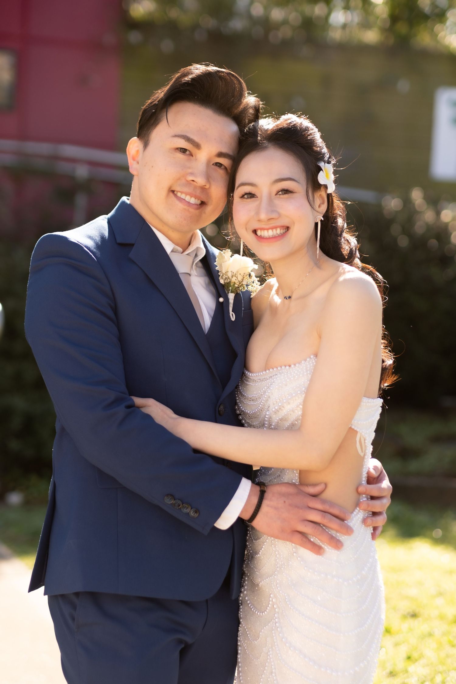 Smiling Asian bride and groom embrace outdoors in sunlight, wearing white wedding dress and navy blue suit.