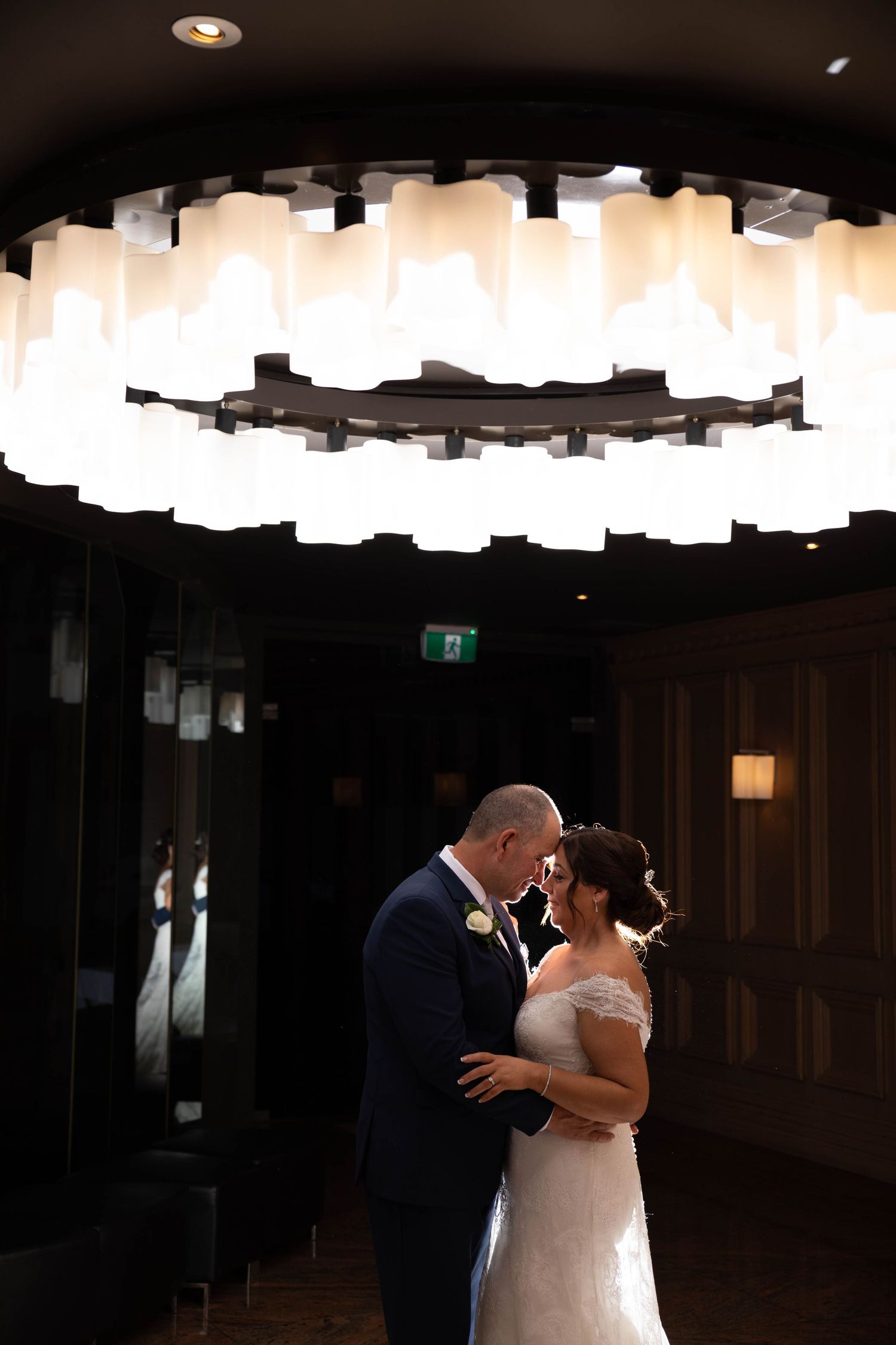 Bride and groom share first dance under a stunning circular chandelier in a dimly lit reception venue.