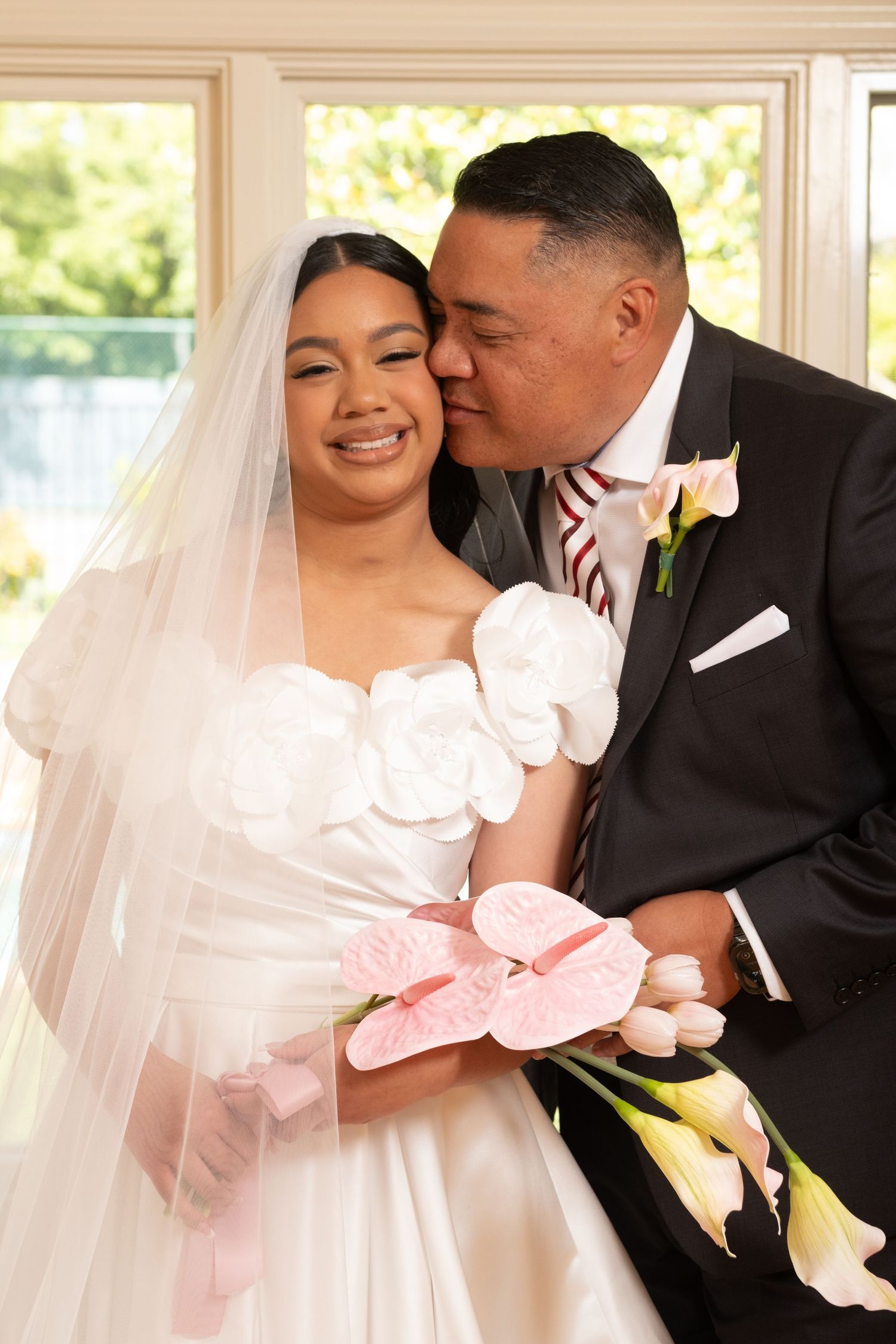 Father kisses daughter bride wearing white floral off-shoulder wedding gown with veil.