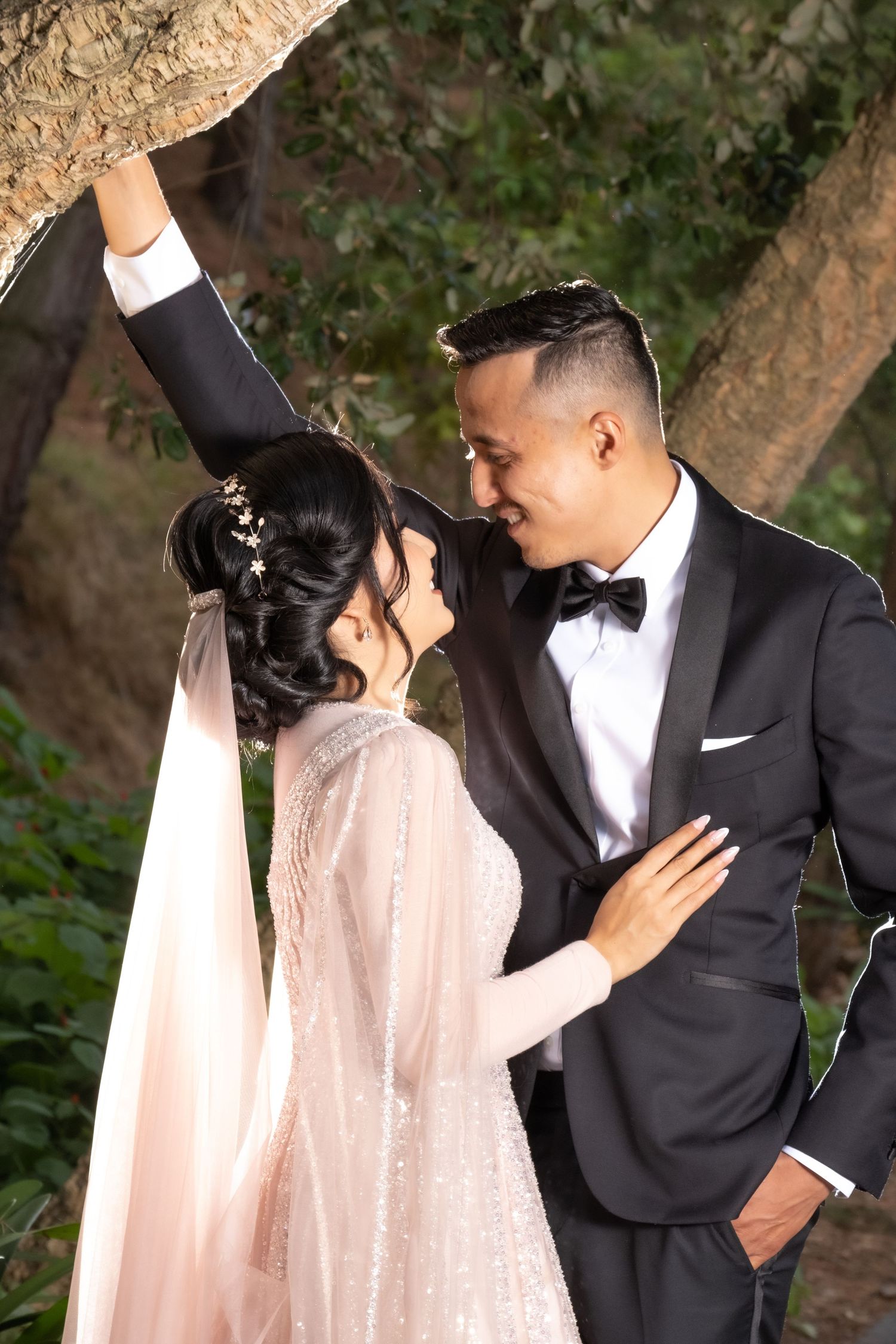 Groom in black tuxedo and bride in sparkly pink gown share romantic moment under a tree outdoors.