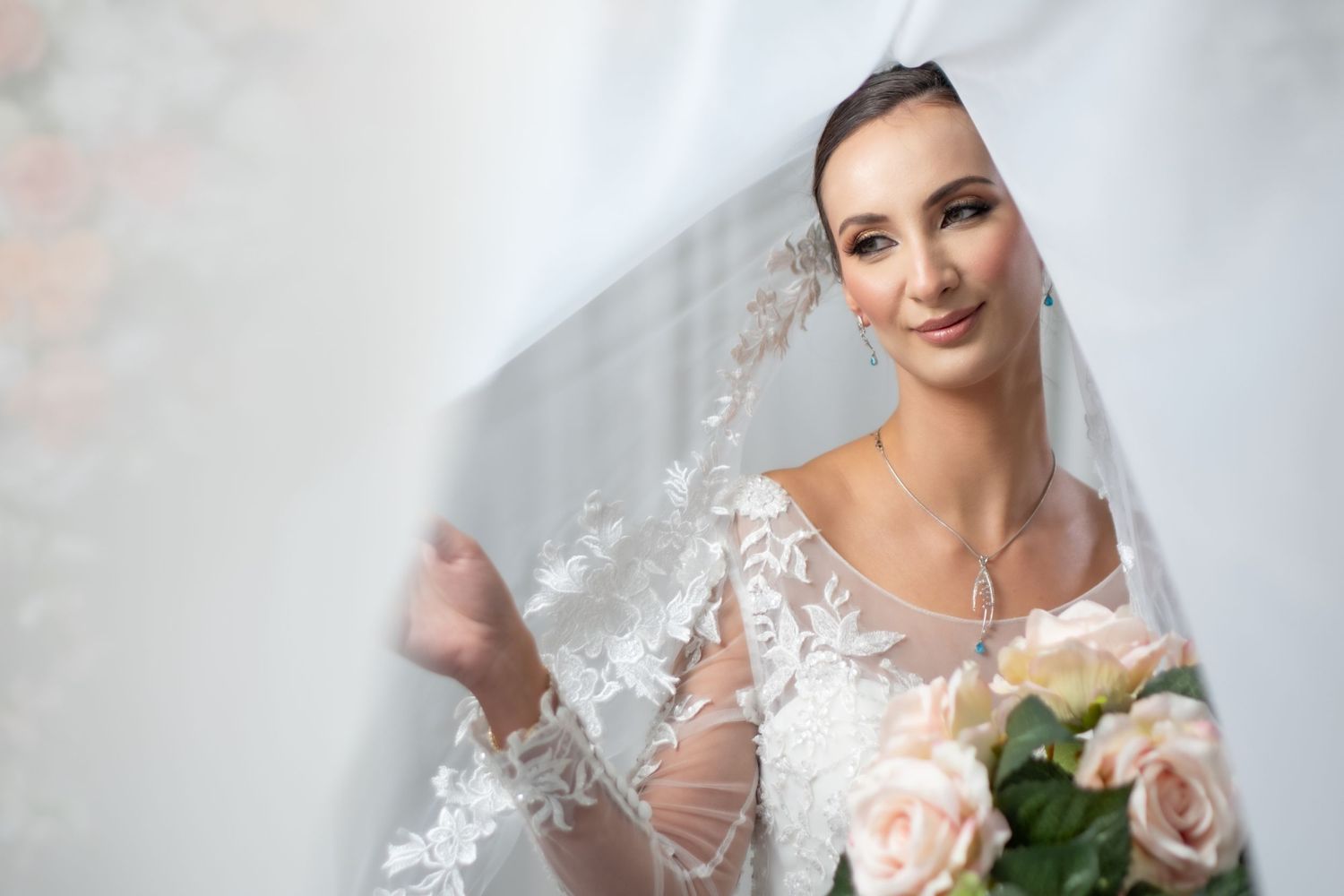 Smiling bride in lace wedding dress holding blush roses under a flowing veil.