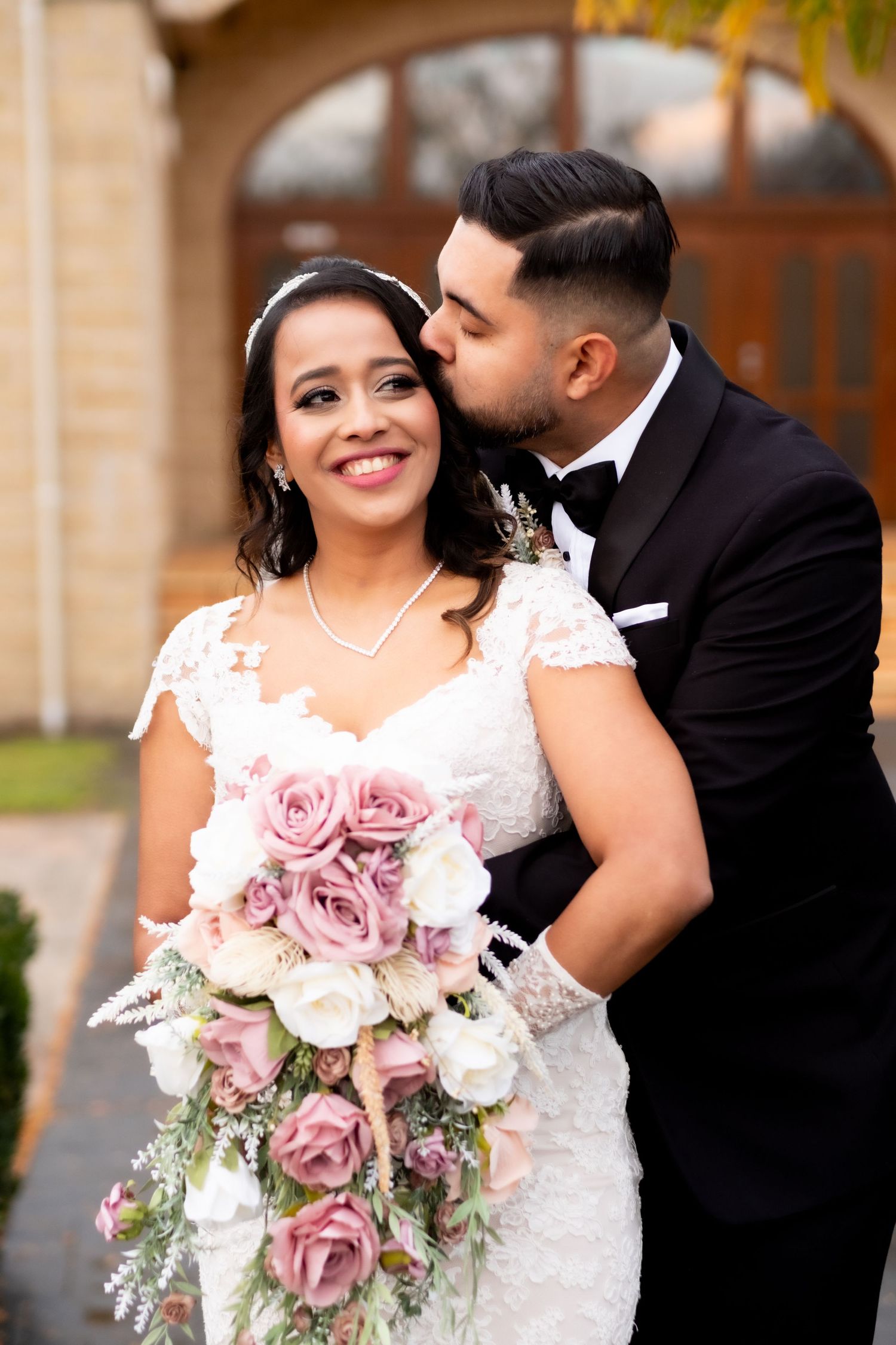 Bride smiling with bouquet of pink roses as groom in black tuxedo kisses her cheek outdoors.