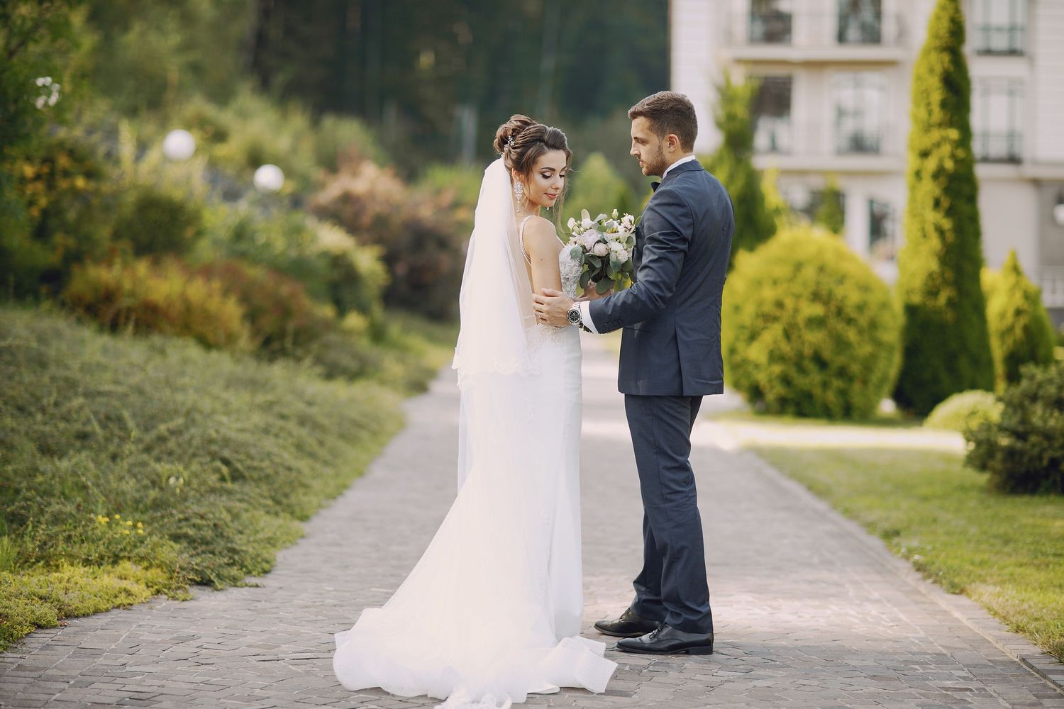 Bride and groom share romantic moment on stone pathway surrounded by lush green gardens.