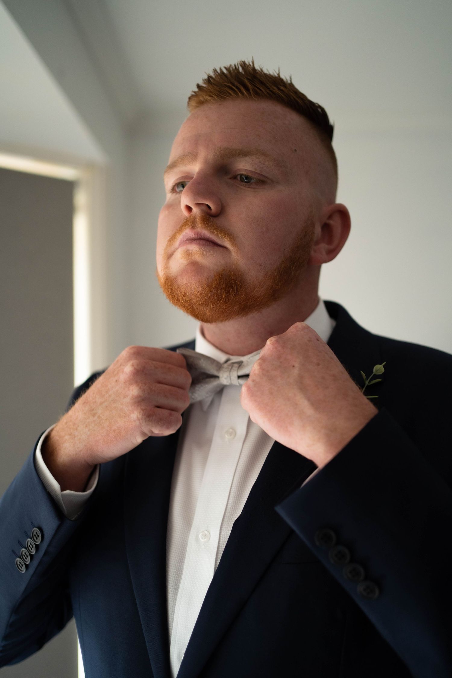 Dapper red-bearded groom in navy suit adjusts bow tie while preparing for his wedding day.