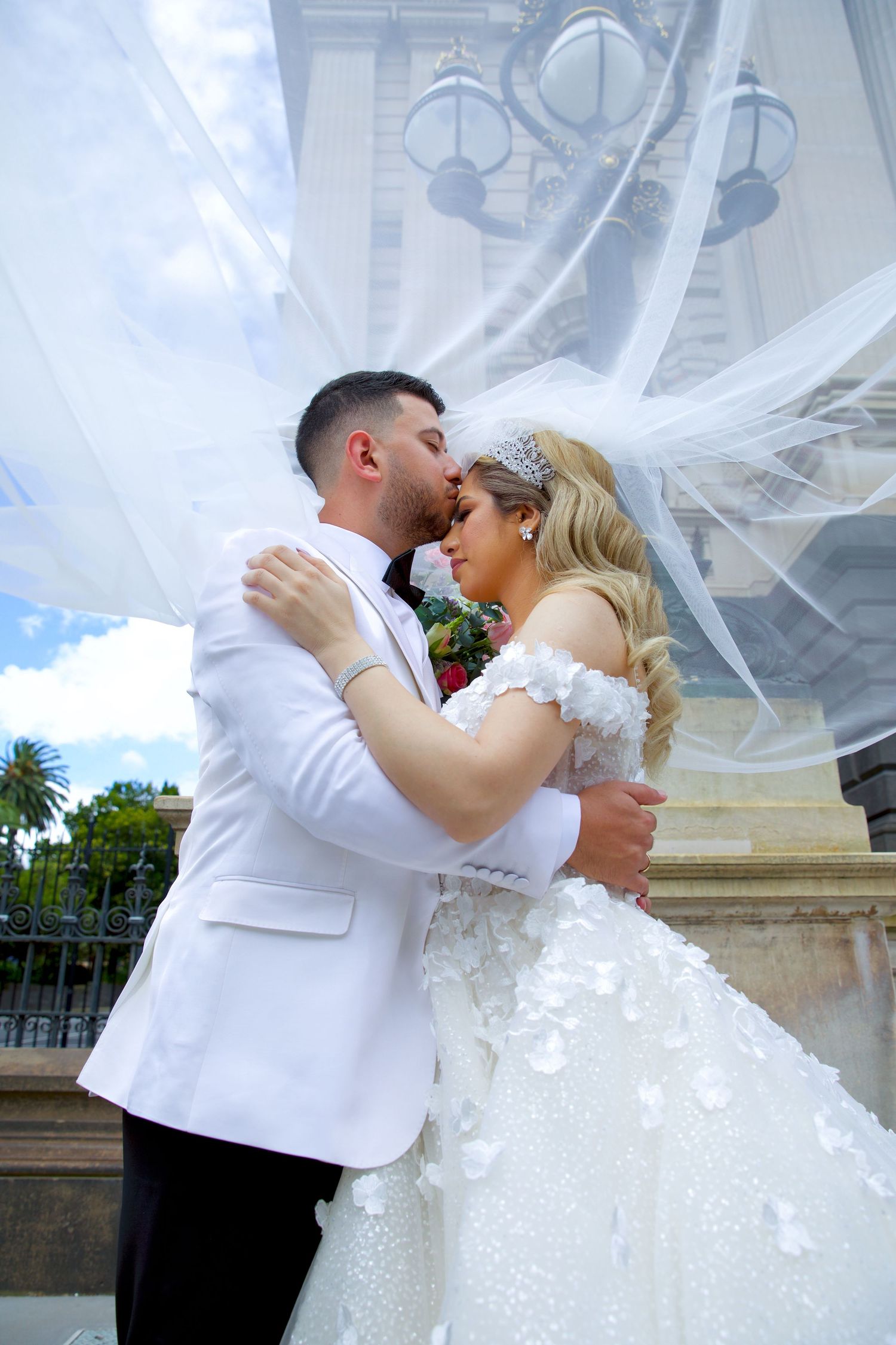 Wedding couple kissing passionately under a billowing veil, groom in white tuxedo, bride in floral ballgown.