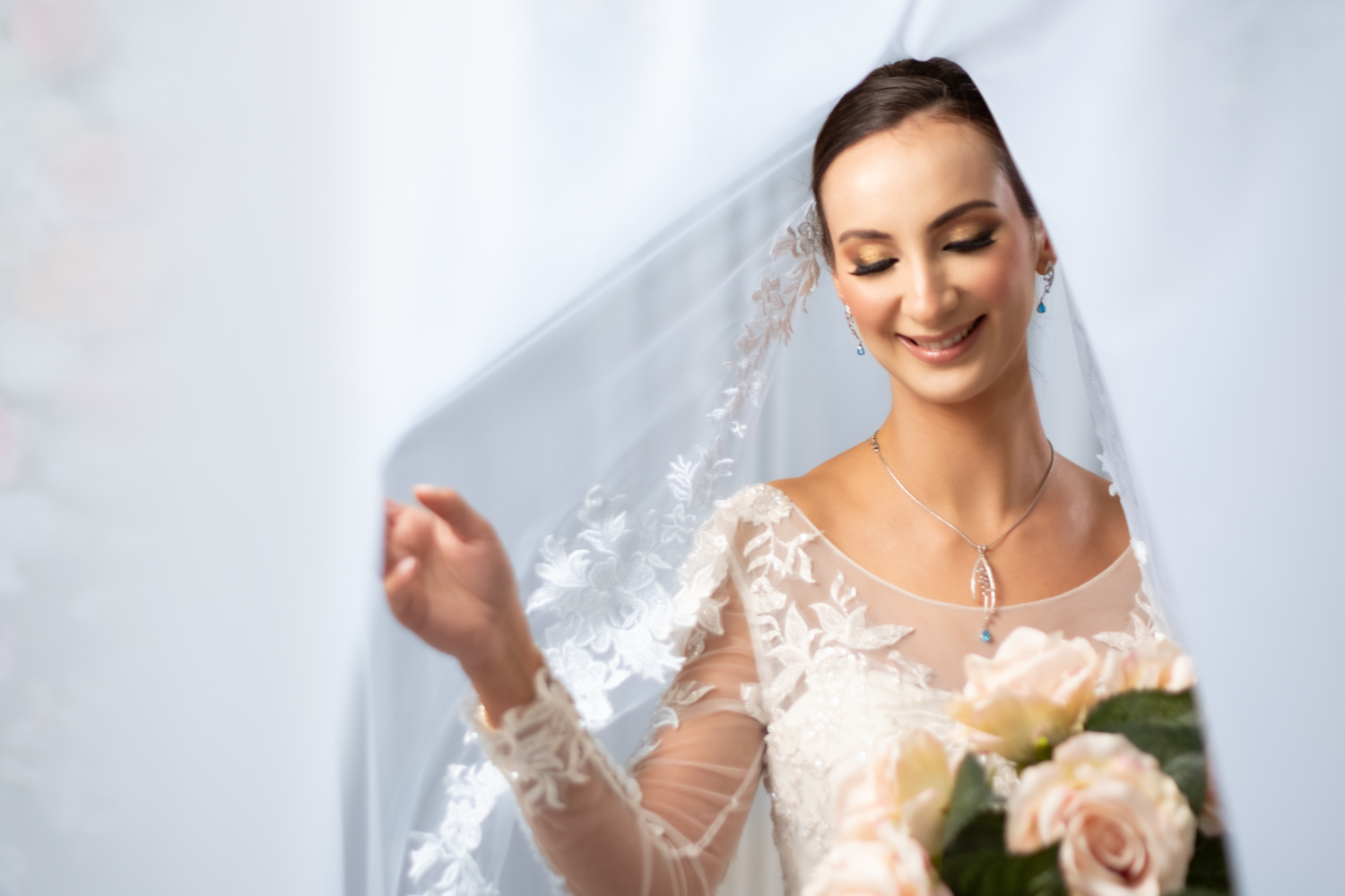 Elegant bride in lace wedding gown smiles softly while holding roses beneath a flowing veil.