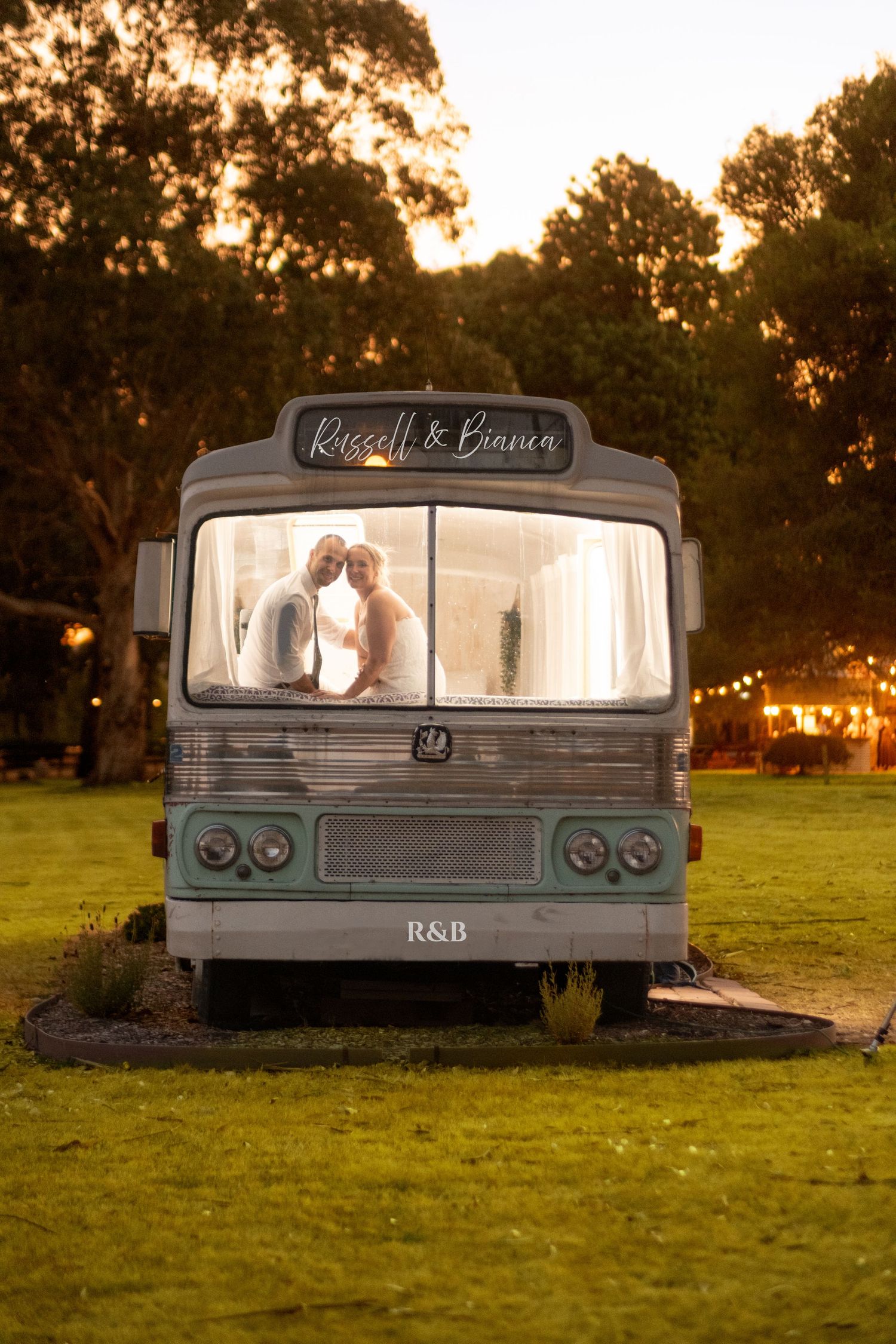 Wedding couple kissing inside a vintage bus decorated with Russell & Bianca signage at golden hour.
