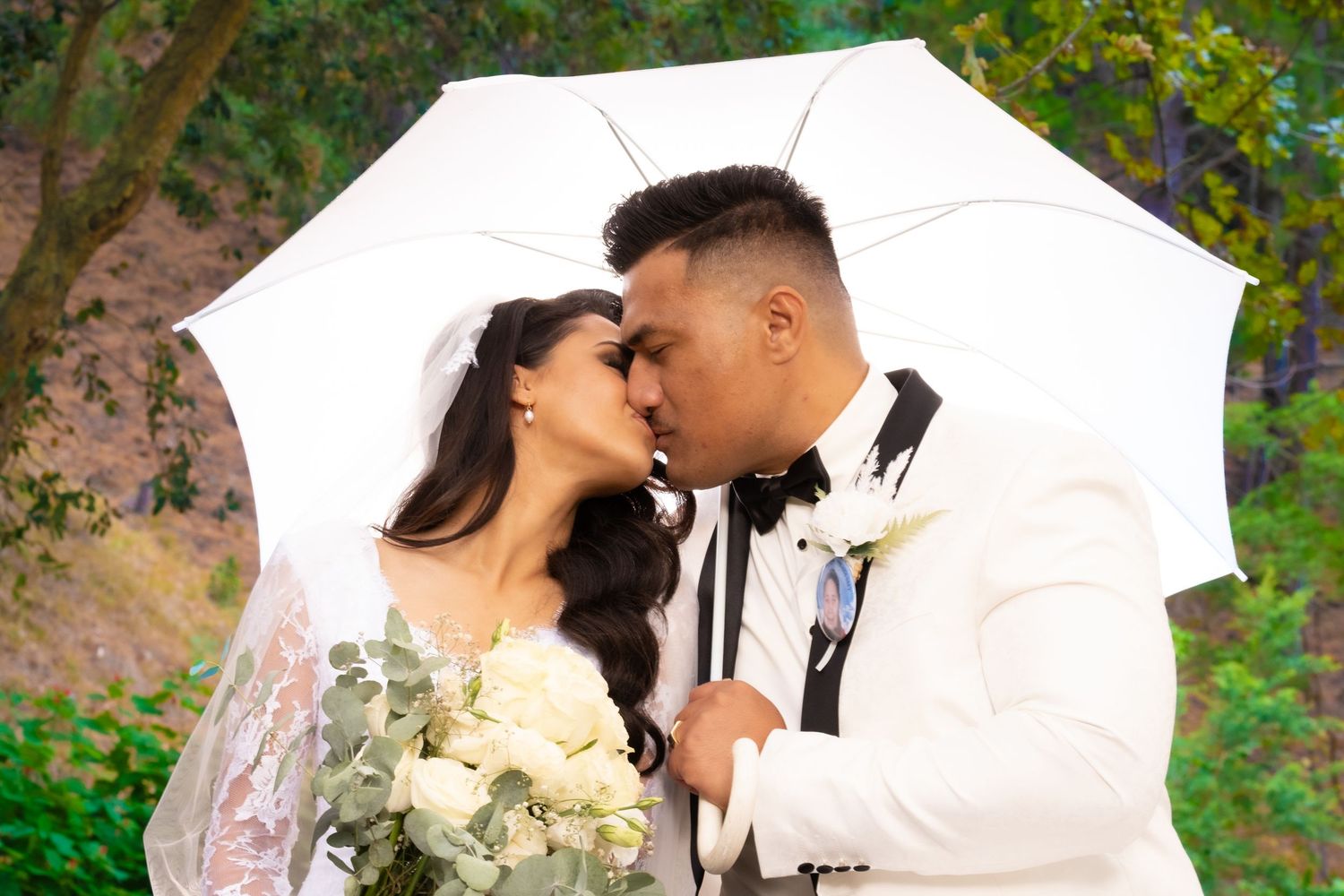 A couple shares a romantic kiss under a white umbrella on their wedding day, bride holding a bouquet of white roses.
