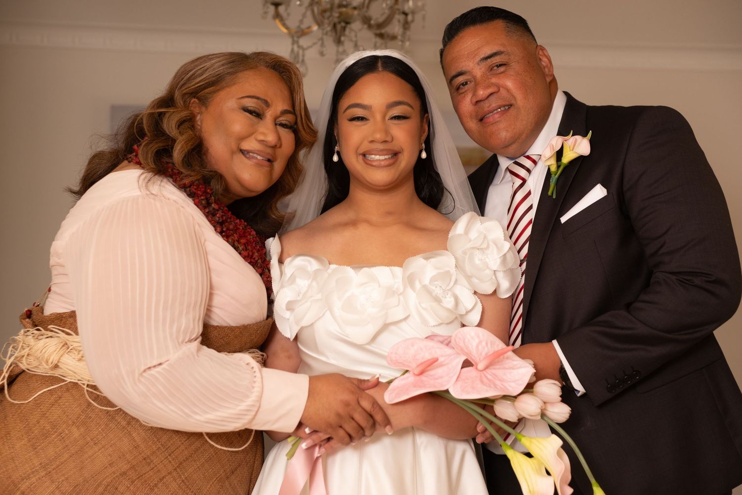 Smiling Polynesian bride poses with parents wearing traditional attire at wedding celebration.