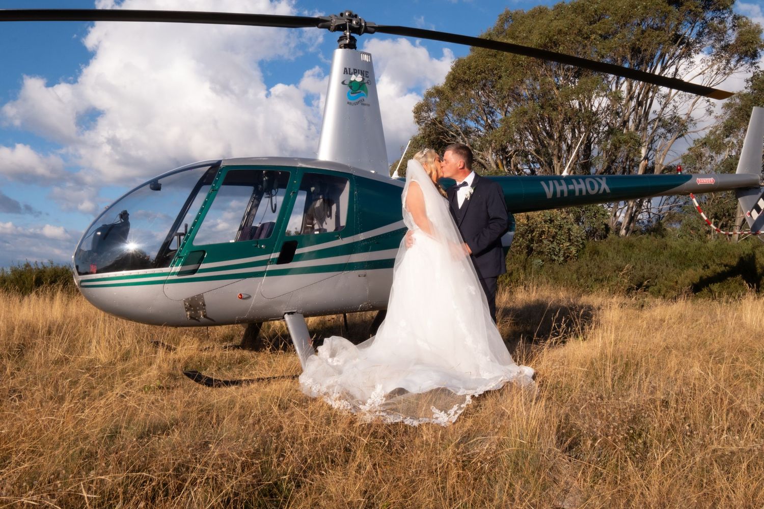 Newlyweds share a kiss beside a helicopter in a golden field on wedding day.