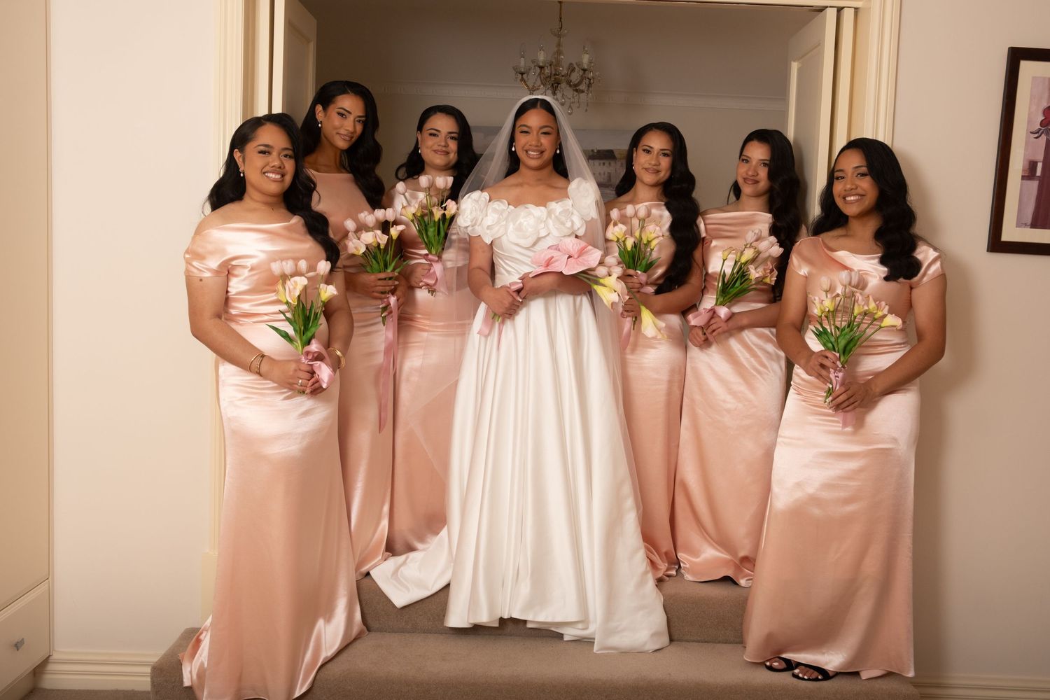 Bride in white gown stands with five bridesmaids in blush satin dresses holding bouquets.