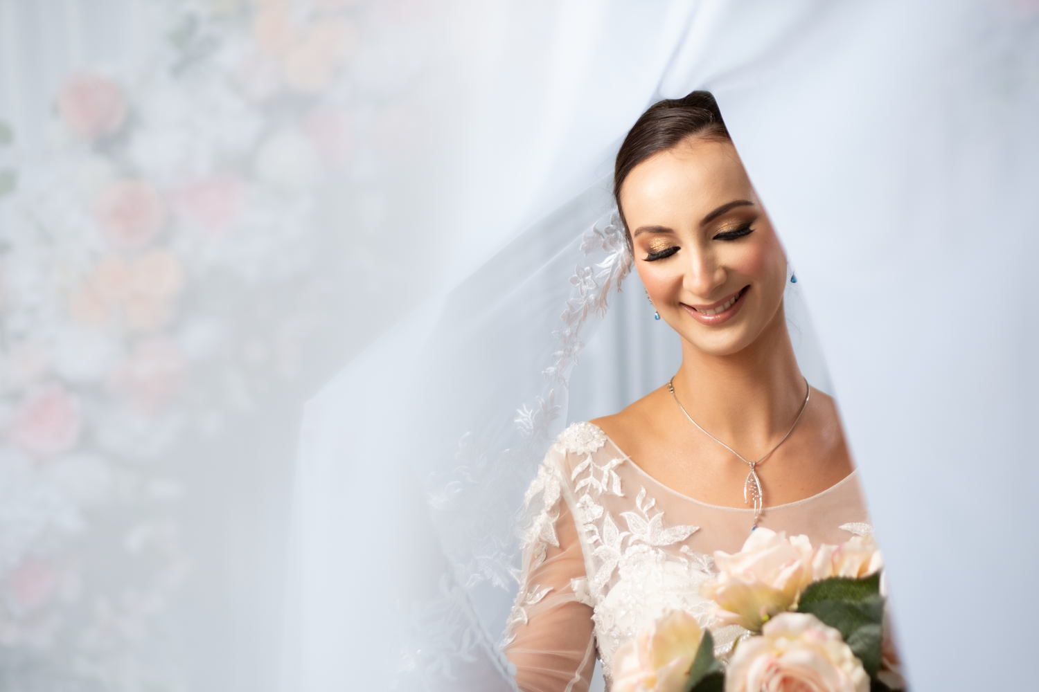 Smiling bride in lace wedding dress holding bouquet of roses under flowing white veil.