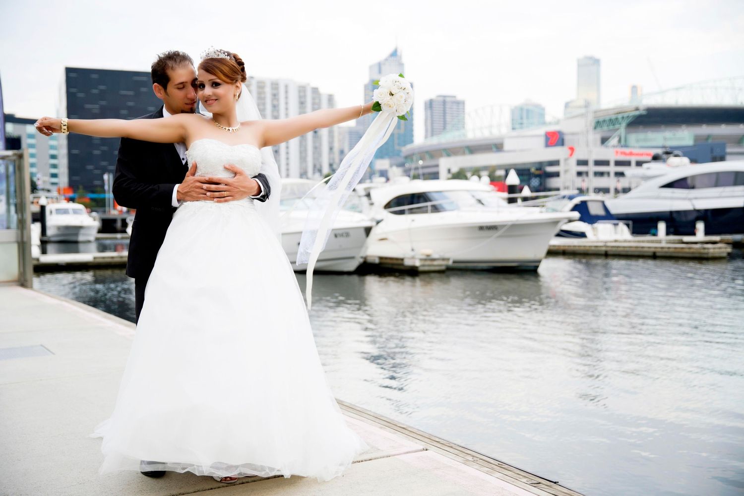 Wedding couple poses at a marina, bride in white gown with arms outstretched, yachts in the background.