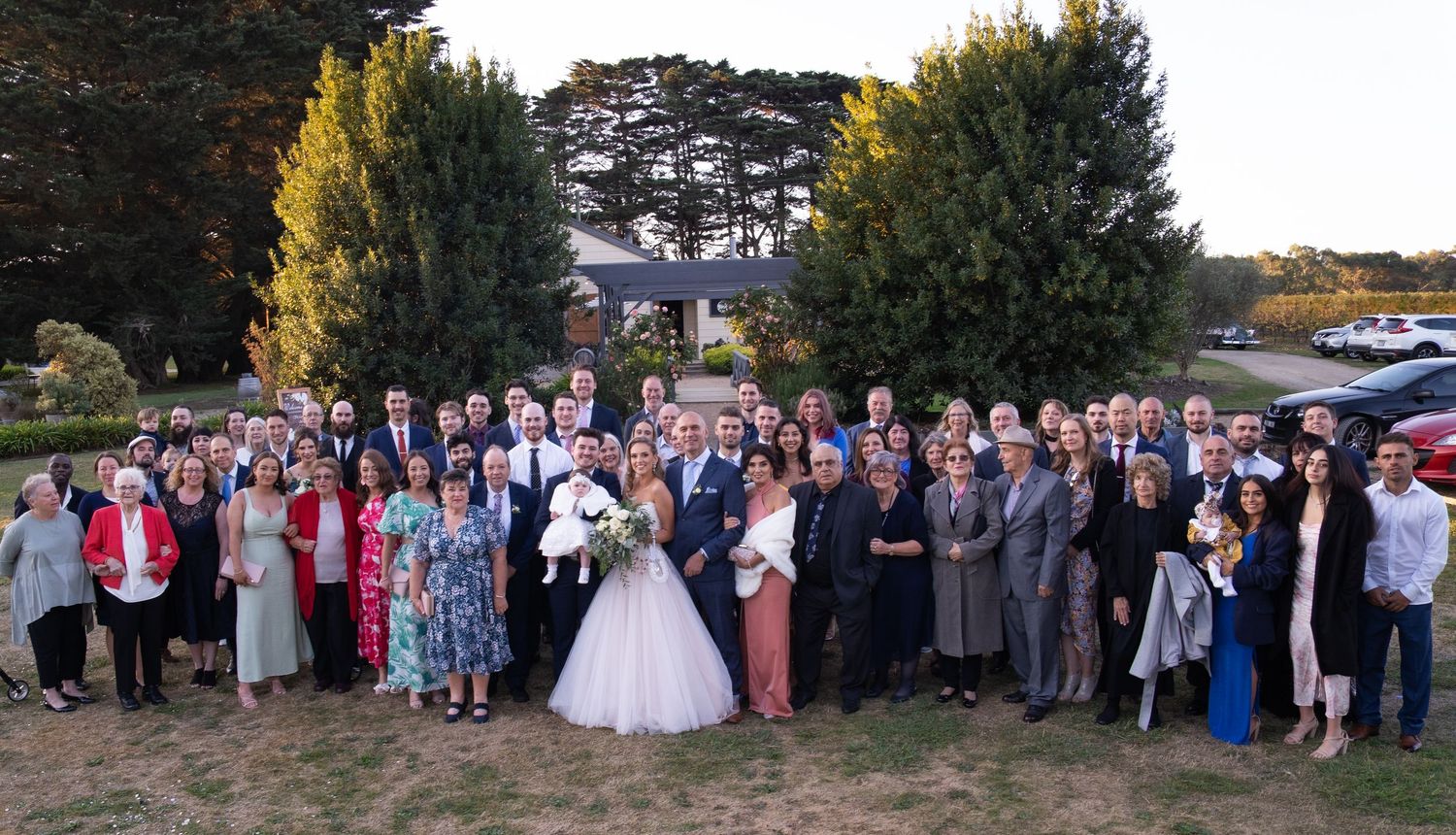 Large wedding party group photo outdoors with bride and groom surrounded by guests among trees.