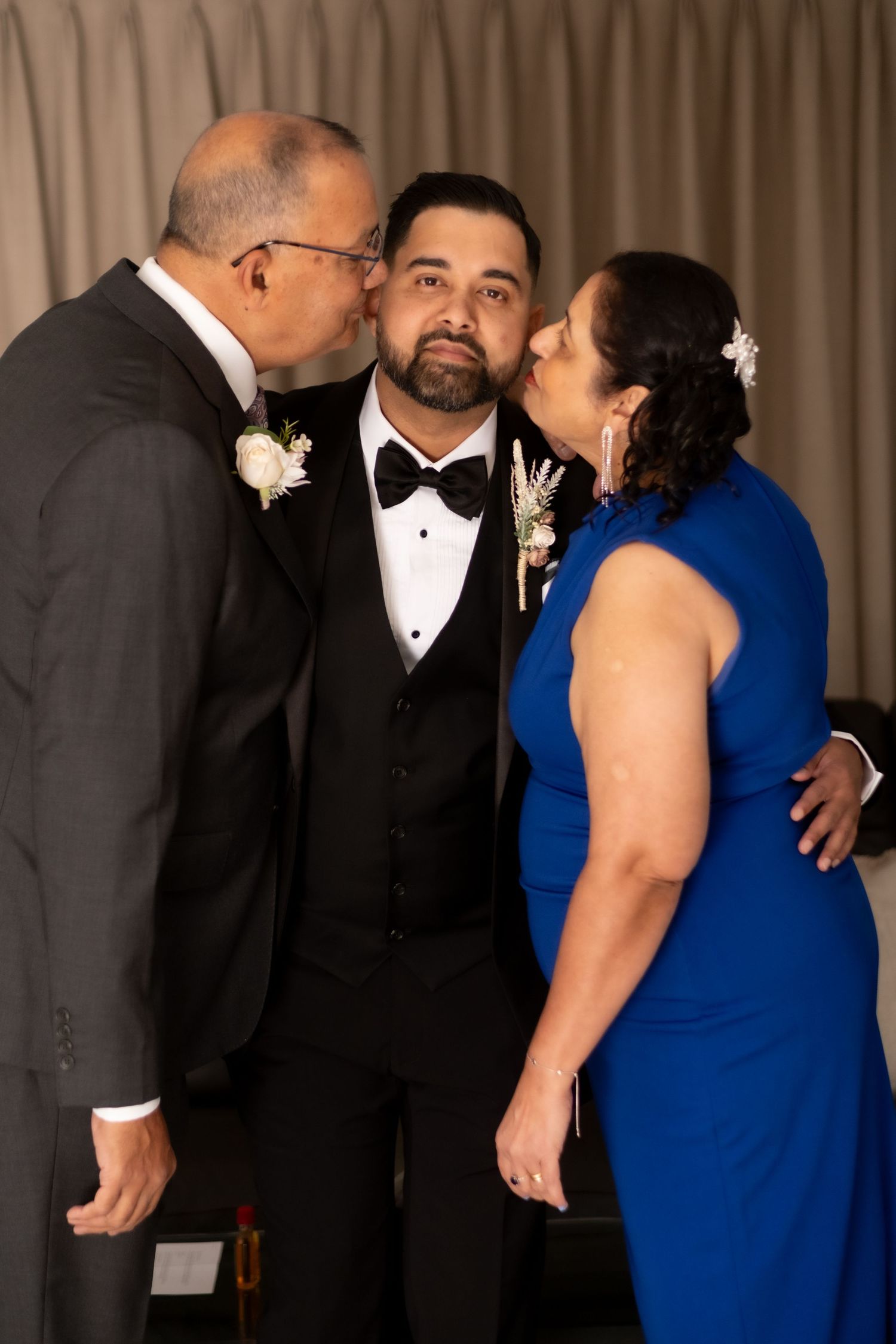 Groom in tuxedo receiving kisses on cheeks from parents, man in gray suit and woman in blue dress, at wedding reception.