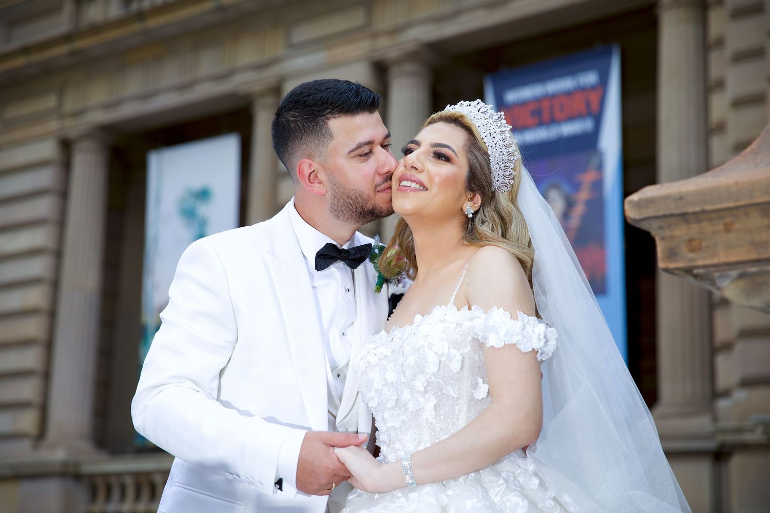 Groom in white tuxedo kisses bride wearing crown and floral off-shoulder gown at wedding venue.