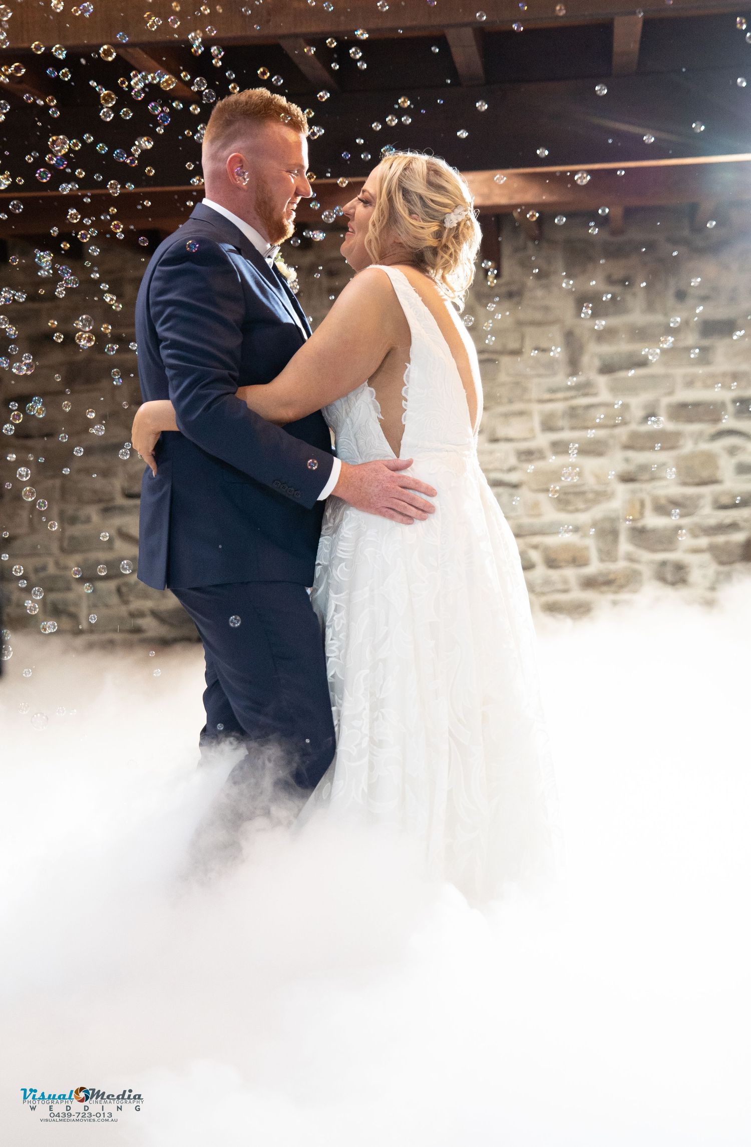 Bride and groom share first dance surrounded by fog and bubbles in rustic barn venue.