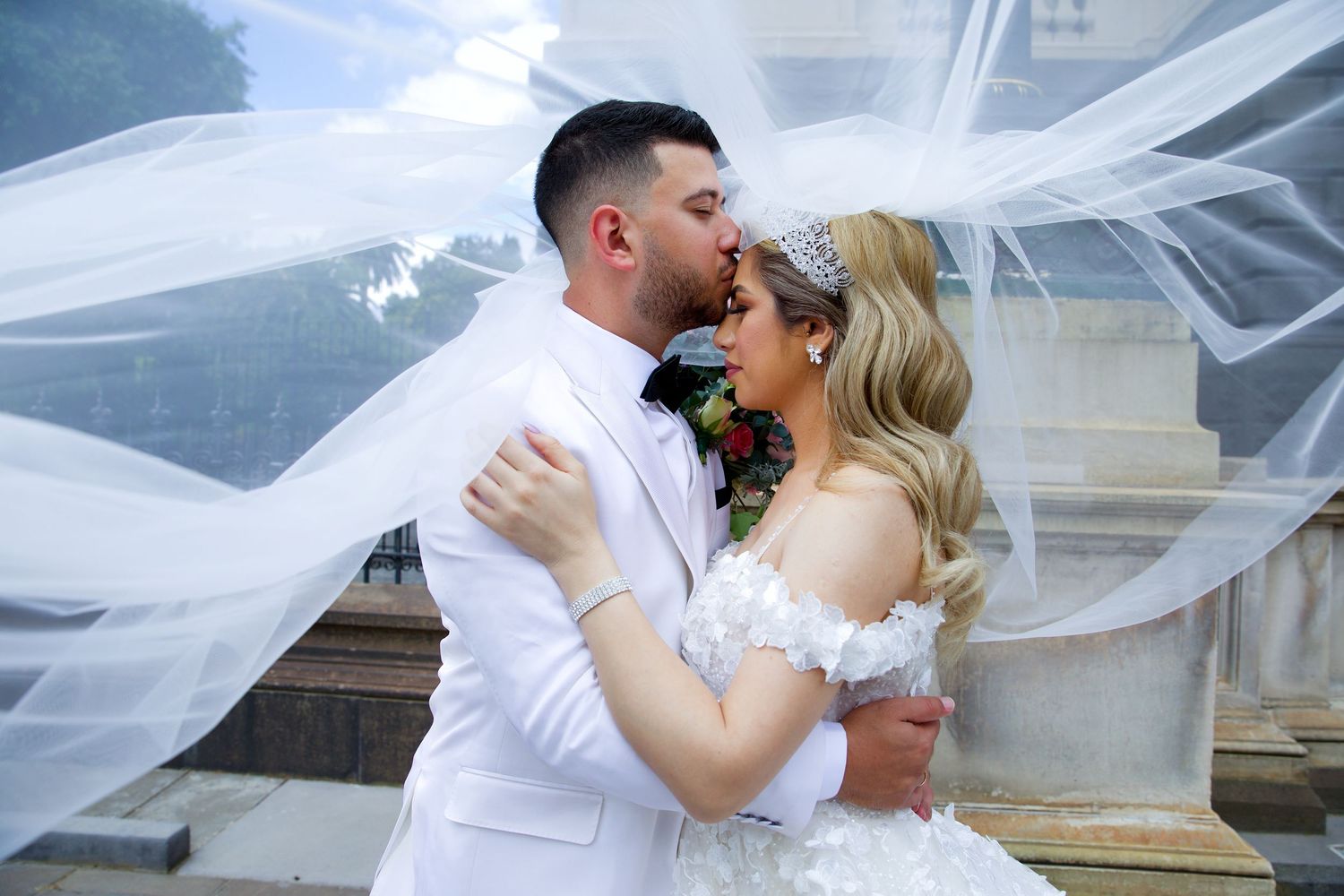 Bride and groom sharing romantic kiss under flowing white veil at outdoor wedding venue.