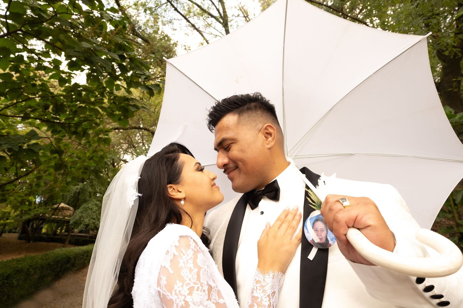 Bride and groom sharing a romantic moment under a white umbrella surrounded by lush green trees.