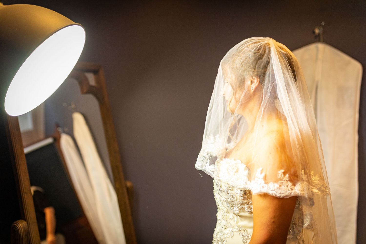 Bride in lace wedding gown standing in soft lamp light beside a mirror before the ceremony.