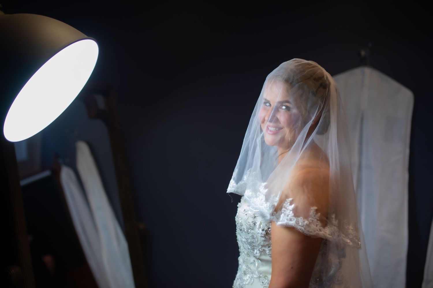 Bride in lace veil and gown smiling softly under studio lighting against a dark background.