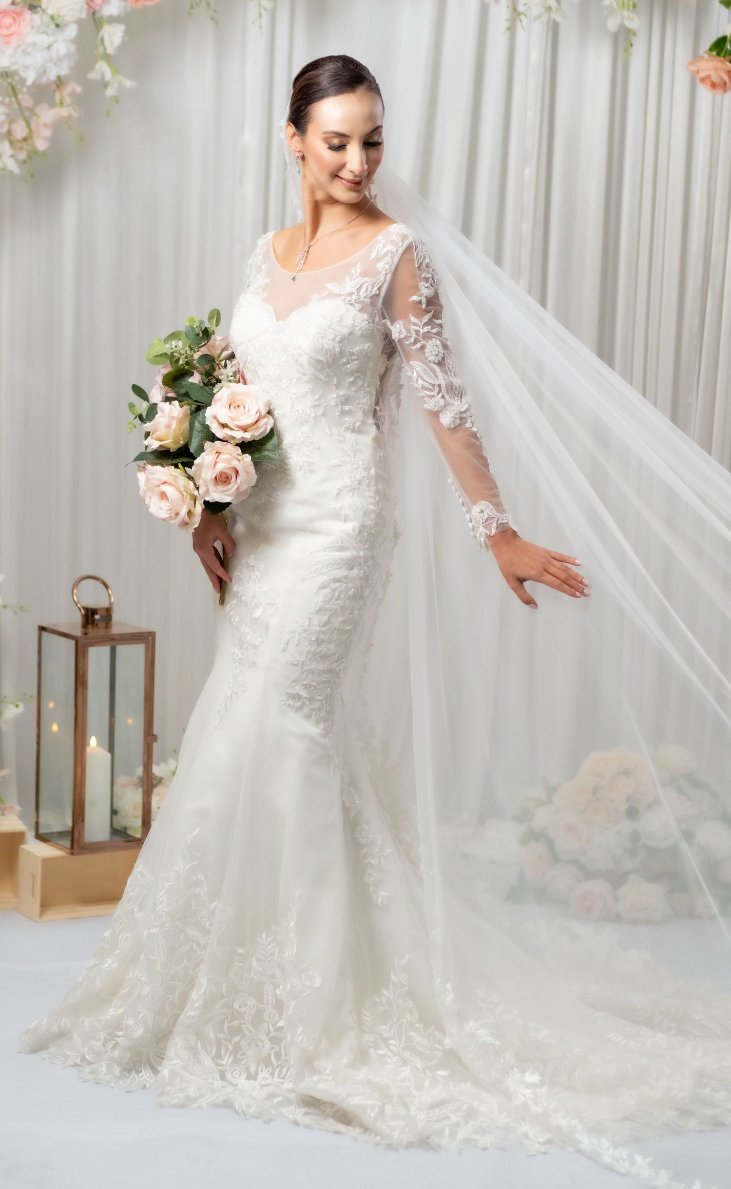Bride in fitted lace mermaid wedding gown with long sleeves holding pink roses bouquet with flowing veil.