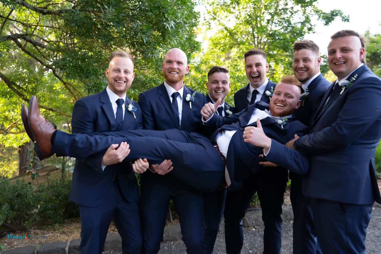 Groomsmen in navy suits playfully lifting groom in outdoor wedding party photo.